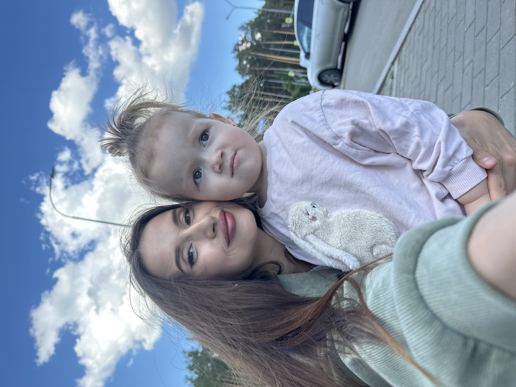 A woman and a young child are in the foreground, with a street and trees in the background. The woman is on the left, holding the child who is on the right. Both are looking towards the camera. The woman has long brown hair and is wearing a green jacket. The child has blonde hair pulled up into a messy bun and is wearing a pink sweatshirt. A small stuffed bunny is attached to the child's sweatshirt. The background shows a paved road and sidewalk, with trees lining the road. A light-colored car is parked on the side of the road. The sky is blue with white clouds. The scene appears to be outdoors, possibly in a park or a residential area.