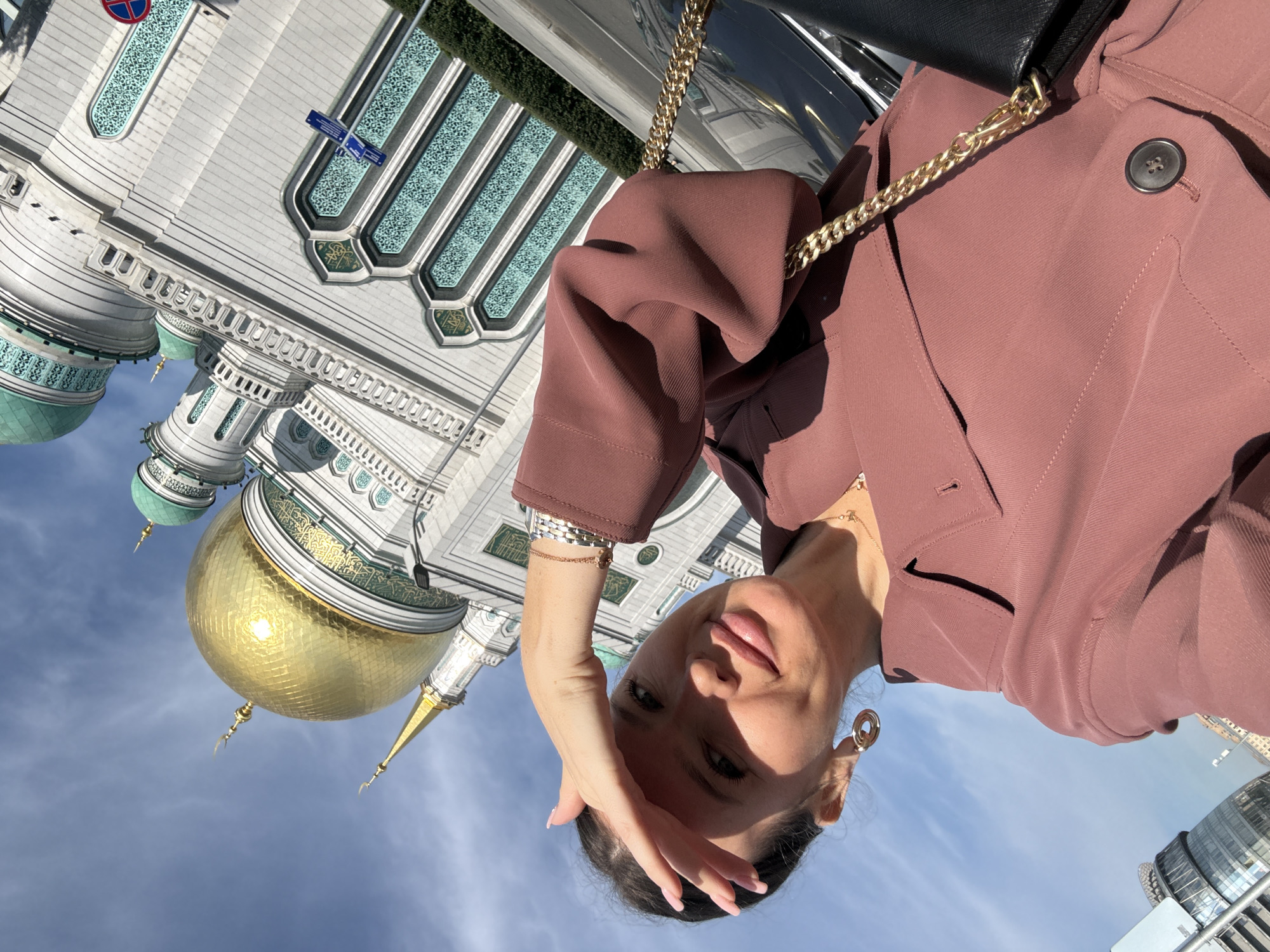 A woman is taking a selfie outdoors in front of the Moscow Cathedral Mosque. The mosque has a large gold dome and intricate white and green architecture. The sky is blue with some clouds. The woman is wearing a reddish-brown jacket and a gold necklace. She has her hand over her forehead, shielding her eyes from the sun. A gold chain handbag is visible over her shoulder.