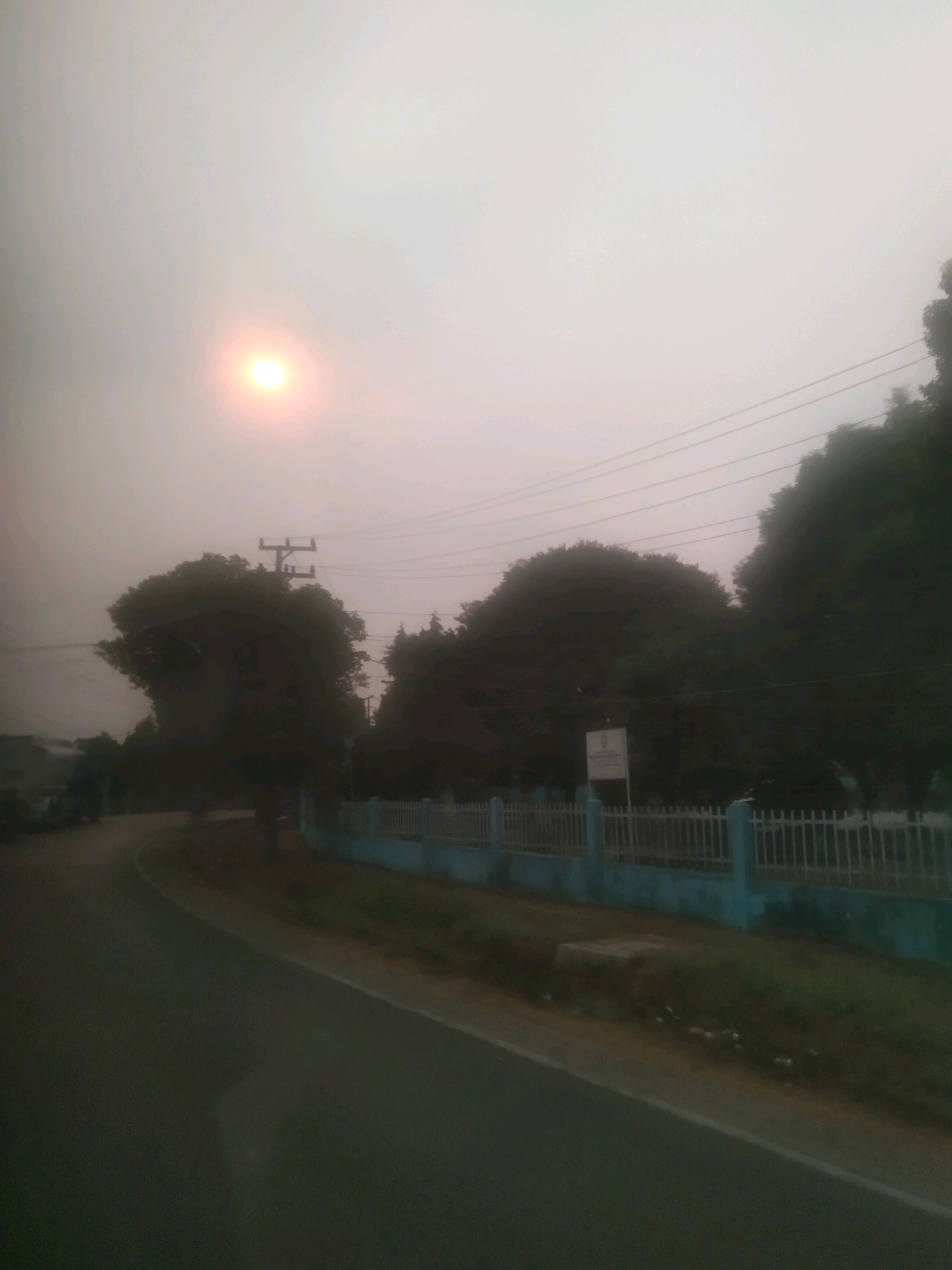 The image depicts a hazy sky with the sun visible as a soft, diffused circle in the upper left quadrant. In the foreground, a paved road curves to the left, appearing to be a street in the city of Tanjung Raja, Indonesia. Along the right side of the road, a light blue fence with white metal railings runs parallel. Behind the fence, dense foliage from trees is visible, with some trees appearing taller than others. A utility pole with power lines stretches across the upper portion of the image. The overall atmosphere suggests a potentially smoky or dusty environment, or perhaps dawn or dusk, given the diffused sunlight. The scene appears to be an outdoor, roadside environment, possibly a suburban or semi-rural area within Tanjung Raja.