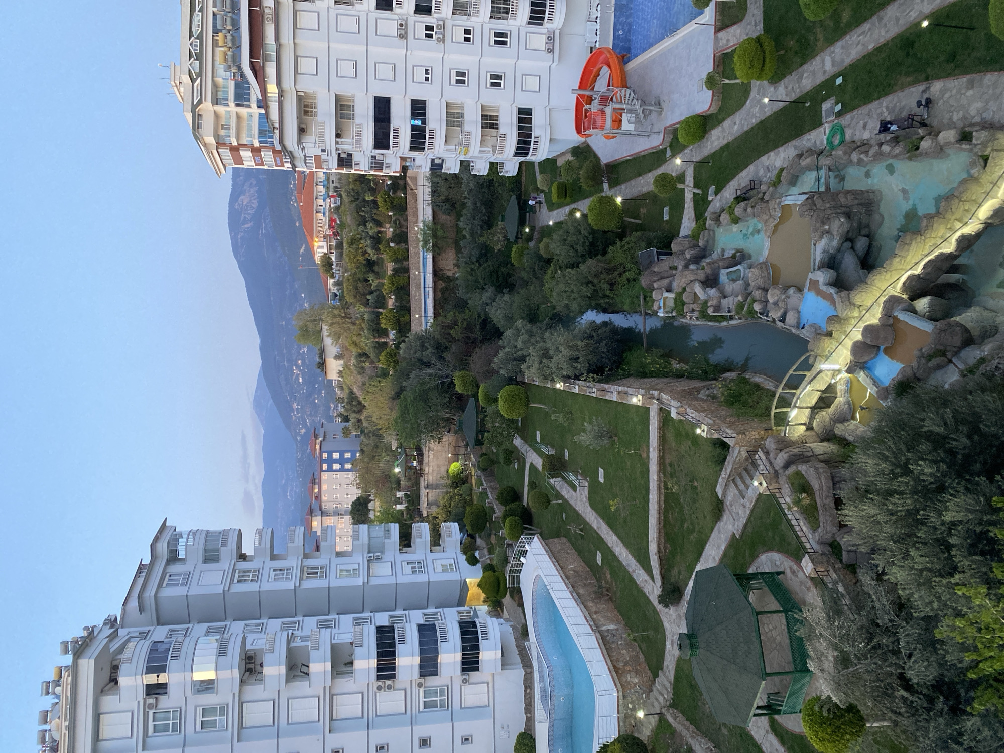 The image shows an elevated view of Alanya, Turkey. In the foreground are two tall, modern apartment buildings with multiple floors and windows. The buildings are white, with gray accents and balconies. Between the two buildings is a landscape with trees, grass, and pools. There is an artificial waterfall with rocks, as well as a pool with an orange water slide. A stone path leads through the grass and trees, and there is a small green gazebo in the bottom right. In the background, behind the buildings and trees, is a mountain range. The sky is light blue and clear.
