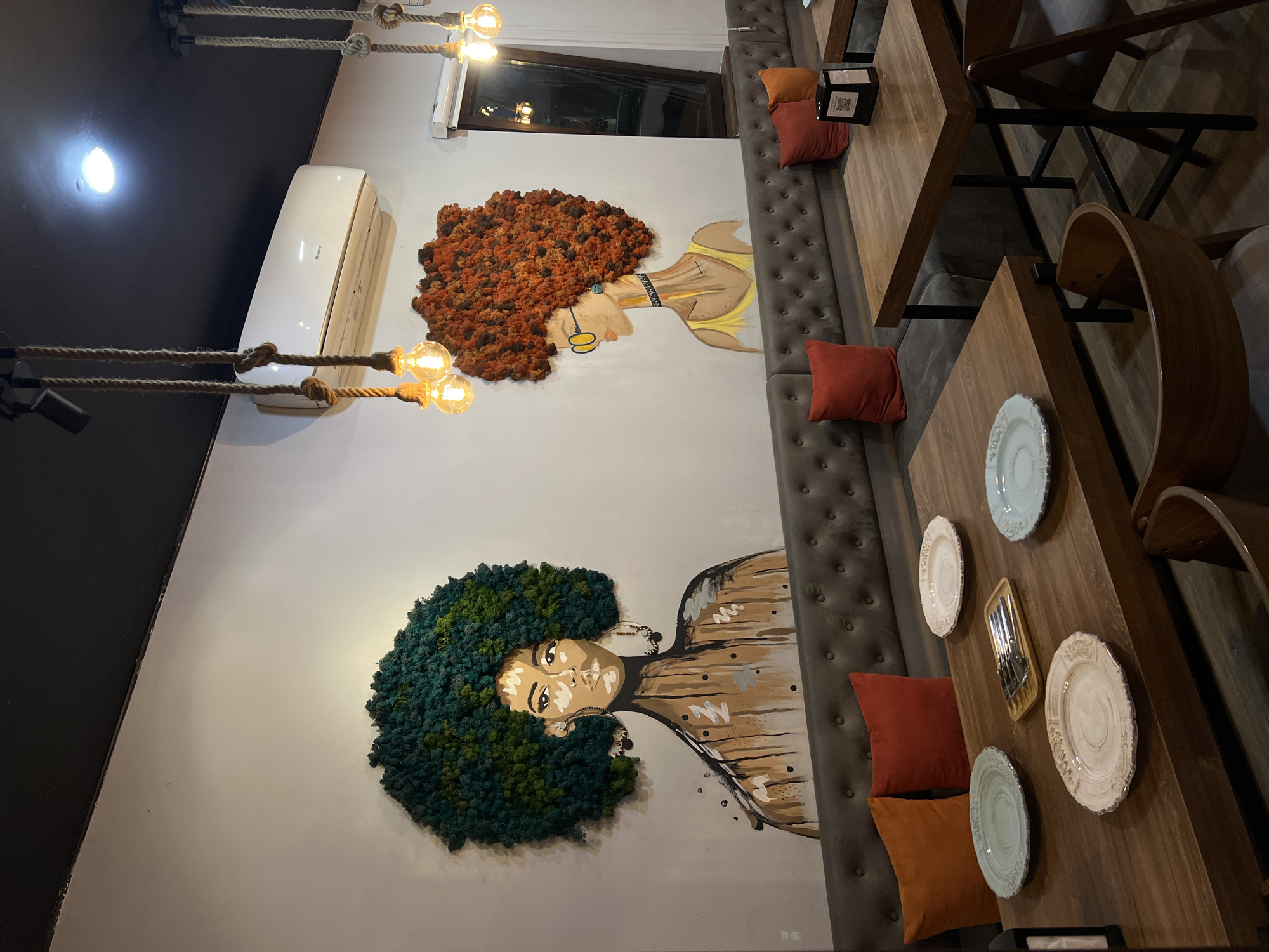 The image captures the interior of a restaurant in Yerevan, Armenia. Two large murals of women with Afro hairstyles adorn the wall above a tufted gray bench. The woman in the top mural has orange hair, gold earrings, and a yellow dress. The woman in the bottom mural has green hair and a brown dress. Tables are set with decorative plates. Light fixtures with rope and bulb lights hang from the ceiling. An air conditioning unit is mounted on the wall above the orange-haired mural. Brown and red pillows are placed against the bench.