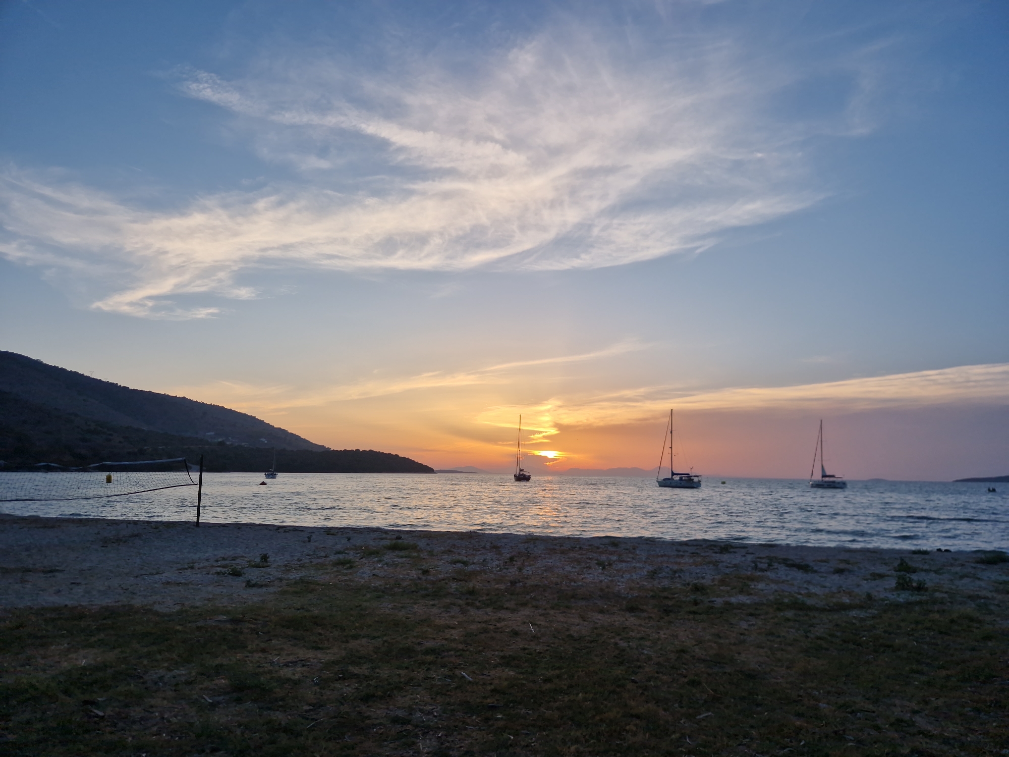 The image captures a serene sunset scene on a beach in Plataria, Greece. The foreground features a sandy and grassy area, with a volleyball net set up near the water's edge. The sea is calm, reflecting the warm hues of the setting sun. Several sailboats are visible on the water, anchored or gently drifting. The background is dominated by a silhouetted hillside covered in vegetation, with the sky displaying soft pastel colors and wispy clouds. The overall atmosphere is peaceful and tranquil.
