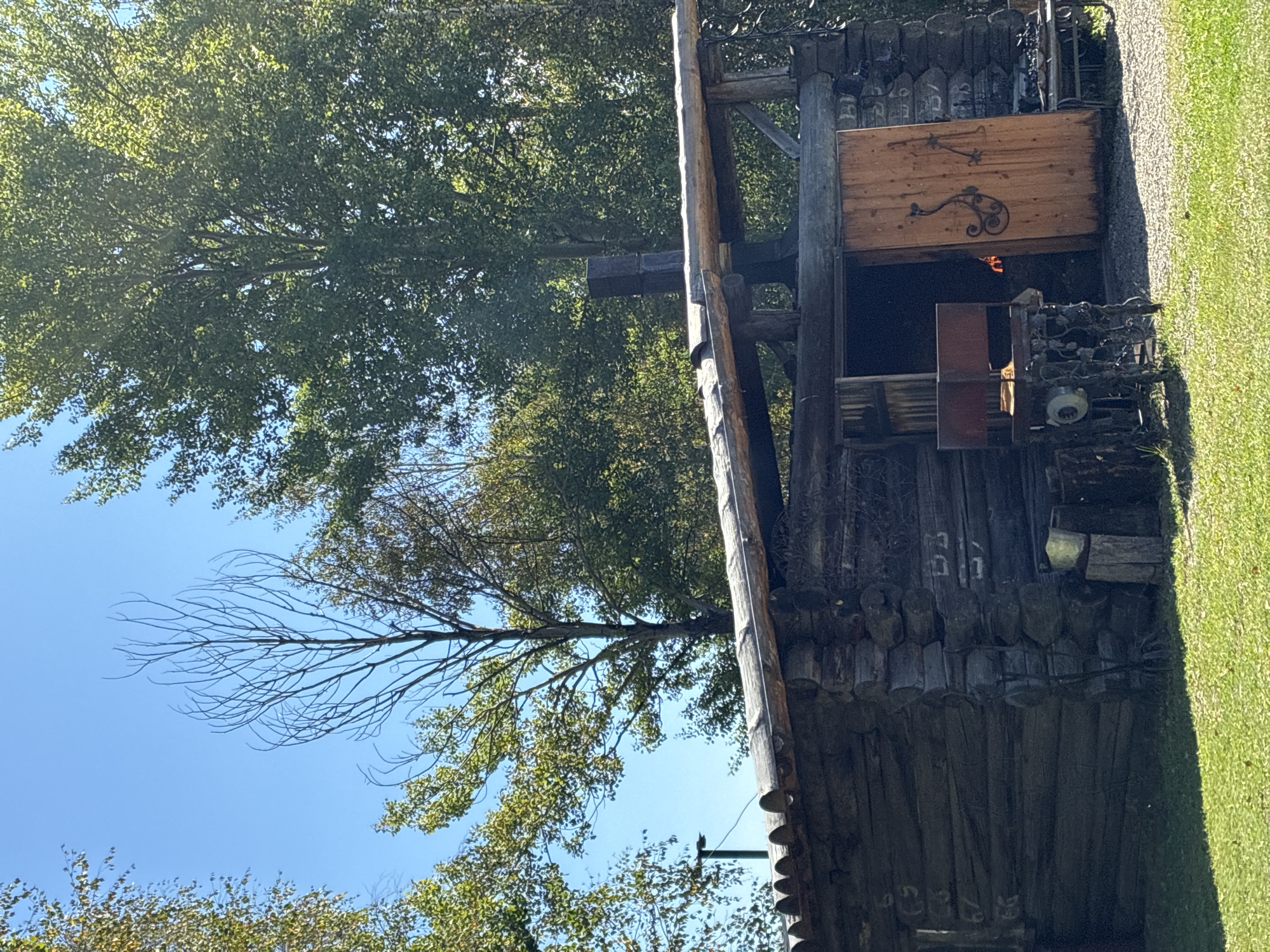 The image depicts an outdoor scene in front of a rustic log building. The building has a wooden roof supported by logs and a large wooden door, which is partially open, revealing a glimpse of fire inside. To the left of the door, there is an old metal contraption, possibly a forge or a piece of machinery. A metal chimney protrudes from the roof of the building. The structure is surrounded by greenery, including grass and trees. The sky is clear and blue. The overall impression is that of a rural or historical setting, possibly a workshop or forge. The image shows no people.
