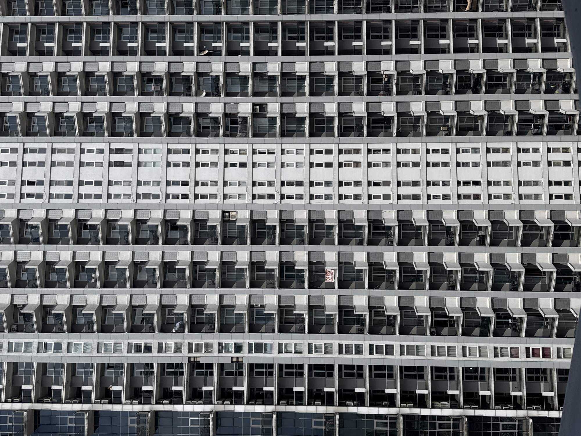 The image shows a full-frame view of a large building facade in Batumi, Georgia. The building features a repetitive pattern of windows and balconies, with horizontal rows alternating between sections with projecting balconies and sections with flat, recessed windows. The color scheme is primarily gray and white, with the balconies and window frames appearing in shades of gray and the recessed sections in white. Some windows reflect the sky or interior spaces, while others are darker. On the balconies, various objects are visible, including furniture, curtains, and other personal belongings. One balcony has a 'FOR RENT' sign displayed. The building is tall, and the perspective is from below, looking upwards.