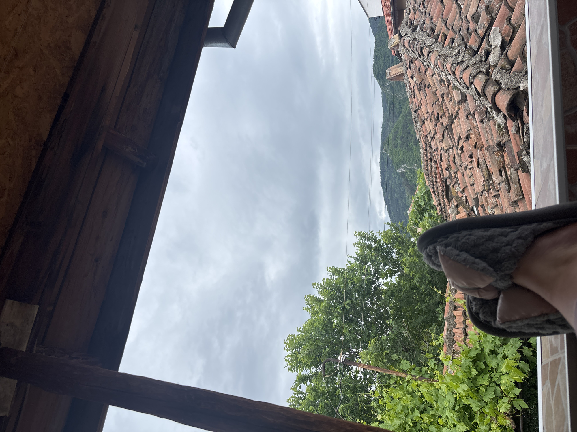 View from an open wooden structure, possibly a window or doorway, overlooking a cloudy sky, lush greenery, and a tiled roof. A mountain is visible in the distance. A slipper is partially visible on the right side of the frame. The location appears to be Asenovgrad, Bulgaria.