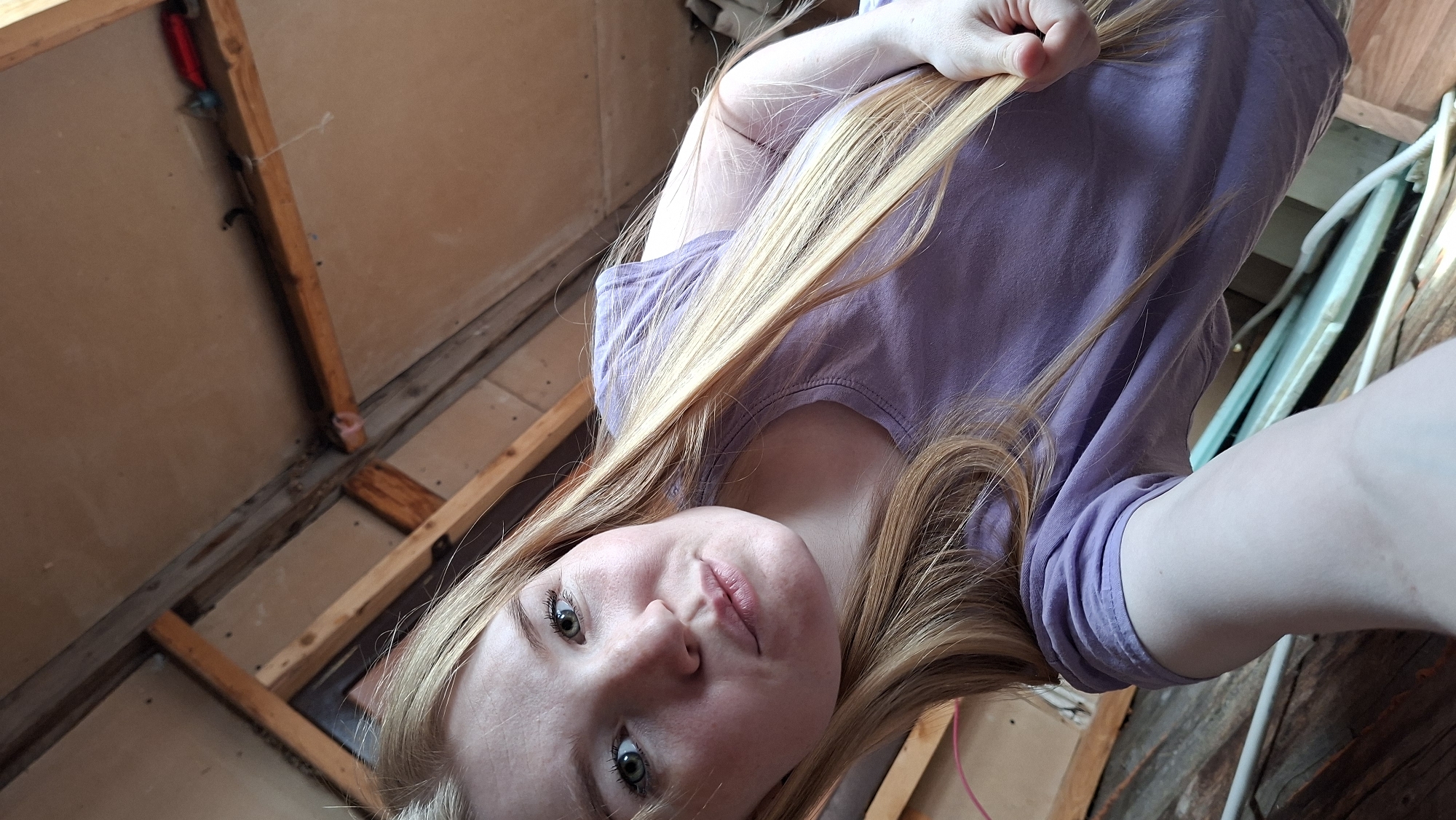 The image is a selfie taken from a low angle, looking up at a young Caucasian female with long blonde hair. She is wearing a light purple t-shirt and has light green eyes. Her lips are slightly pursed. She is holding a section of her hair in her right hand. The background appears to be an unfinished wooden structure, possibly an attic or a construction site, with exposed beams and plywood. There are some wires and pipes visible in the lower part of the frame. The lighting is soft and natural. No specific location in the city of Bolshoy Bukor, Russia, can be identified from the visual information provided.