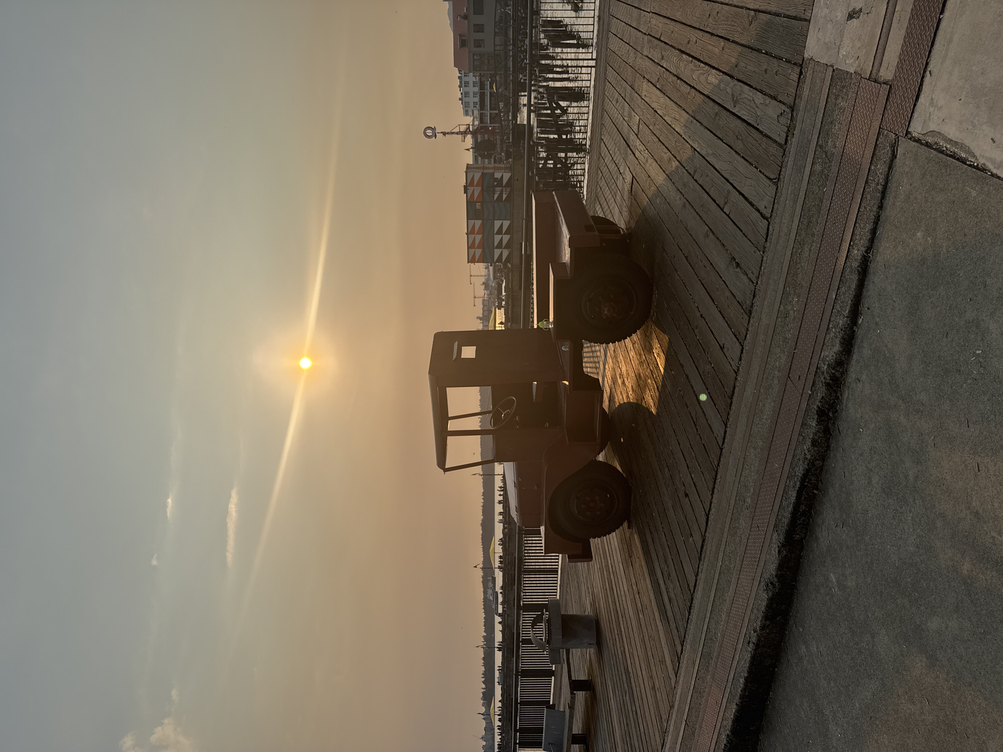 The image depicts a red flatbed truck parked on a wooden pier at a waterfront location in North Vancouver, Canada. The sun is low in the sky, casting a warm, hazy glow and long shadows across the wooden planks. In the background, there is a body of water, a pier with a railing, and buildings, including a structure with a distinct geometric pattern. People are visible on the distant pier, some beneath yellow umbrellas. The sky is a pale orange and yellow, with a few wispy clouds and a visible sun flare. A bench and a trash receptacle are situated to the left of the truck on the pier. The overall scene suggests a late afternoon or early evening setting.