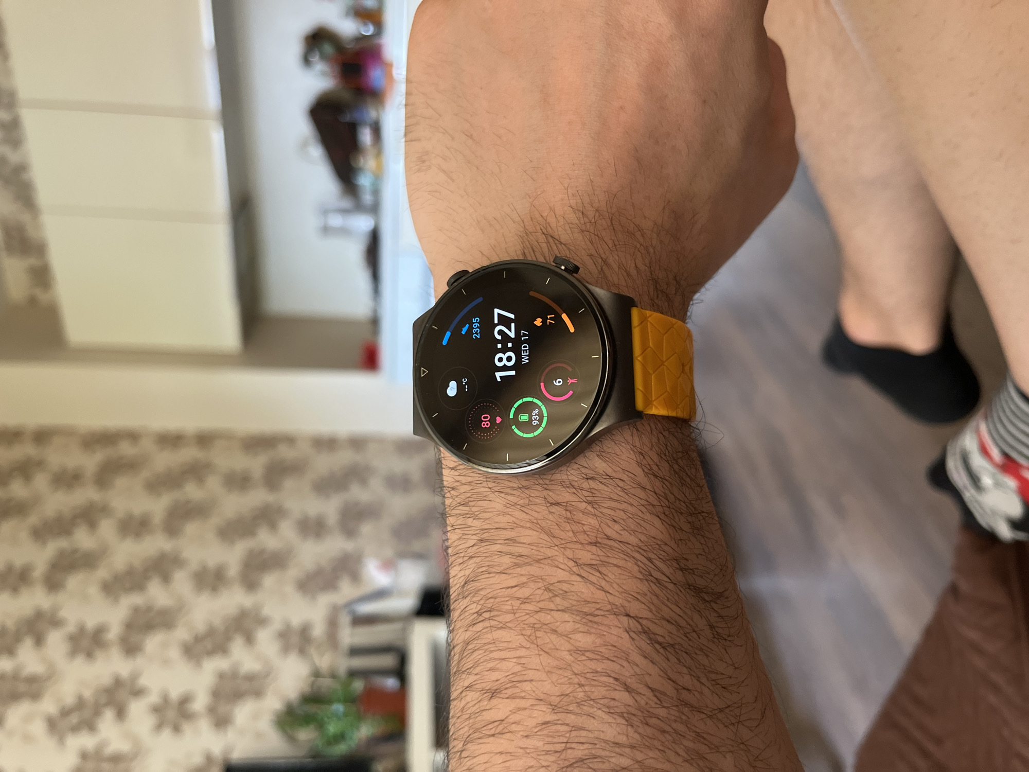 A person's arm, covered in dark hair, wears a smartwatch with a dark gray bezel and a bright yellow patterned strap. The watch face displays the time as 18:27, the date as WED 17, and several metrics including heart rate (80), battery (2395), and activity (93% and 6). The background is blurred, showing a wall with floral wallpaper, kitchen cabinets, and furniture. The person appears to be indoors, possibly in a home environment in Sofia, Bulgaria.