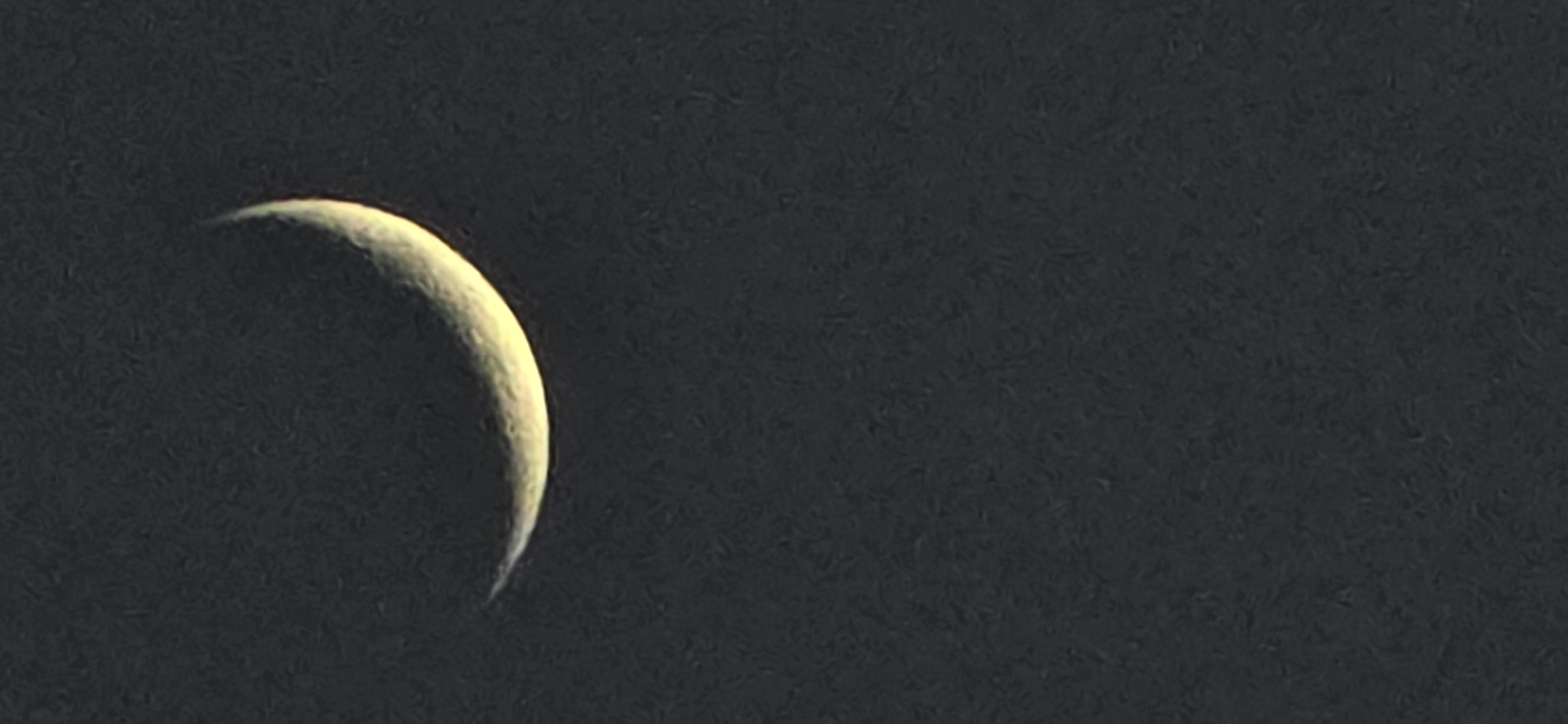 The image shows a crescent moon against a dark sky. The lit portion of the moon is on the left side, with a yellow-white hue. The rest of the sky appears almost black and grainy. No other objects or people are visible. The location of Benasque, Spain is not directly identifiable from this image alone, as it only shows the moon and the sky.
