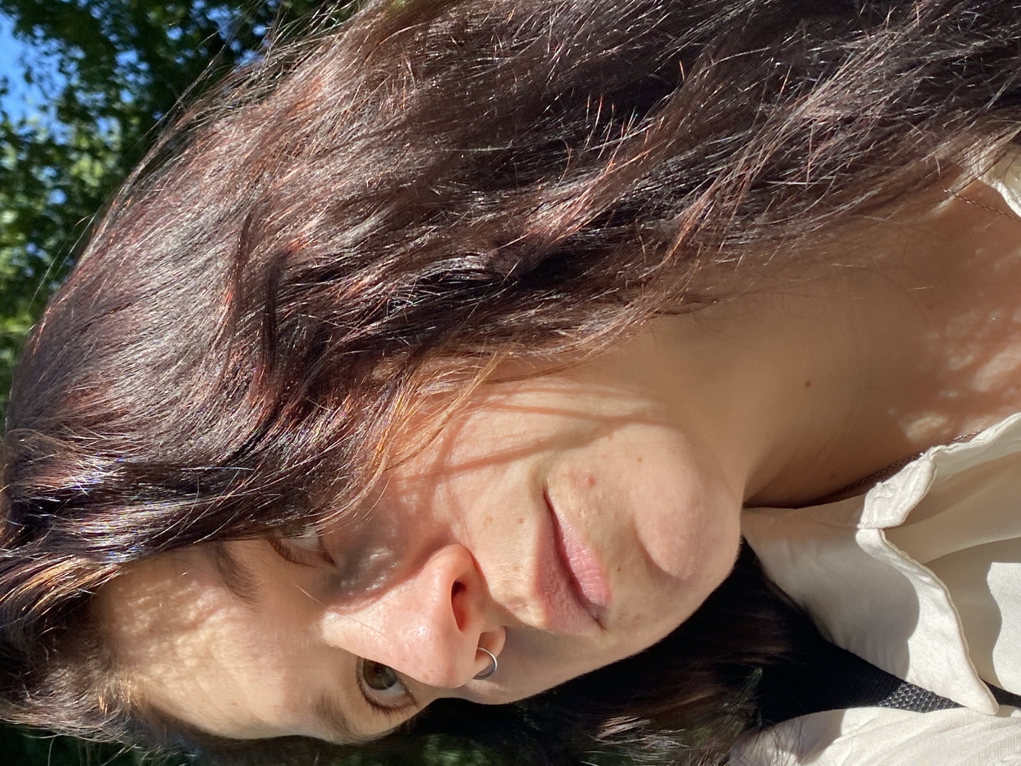 A close-up, eye-level, outdoor medium shot shows a woman with brown hair and fair skin, wearing a white collared shirt. The woman is looking slightly to her left, with a nose ring visible on her left nostril. Sunlight casts dappled shadows across her face, highlighting freckles on her cheeks and nose. The background is blurred, showing green foliage and a bright blue sky, suggestive of a daytime setting. The image does not contain any information about the city of Chaikovsky, Russia.