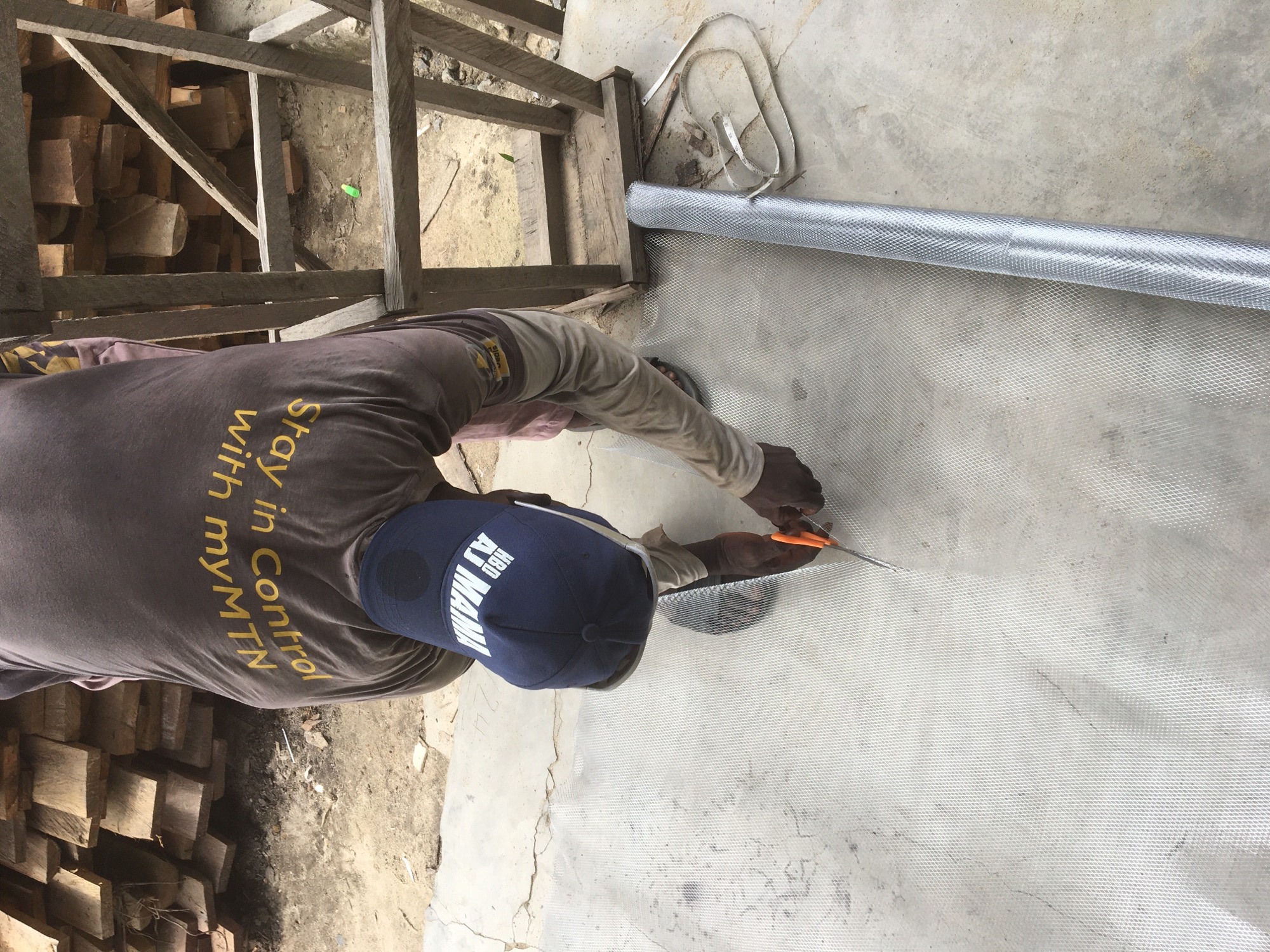 An overhead view captures a person intently cutting a roll of metal mesh on a concrete surface. The image is oriented with the person's body positioned horizontally across the frame, facing towards the top-right.
The individual is wearing a dark brown or grey t-shirt featuring yellow text that reads "Stay in Control with myMTN." Their arms are covered with lighter-colored sleeves, and they are wearing a dark blue baseball cap with white text, "HBD AJ MAMA." They are using orange-handled shears or snips to precisely cut the silvery, fine-patterned metal mesh, which is unrolled from a larger spool. A white fabric measuring tape is also visible near the rolled mesh on the concrete.
In the background, to the left of the person, there is a rustic wooden structure, possibly scaffolding or part of a building frame. Behind this structure, a stack of rough-cut wooden blocks or logs is visible. The ground beneath the wooden structure is dirt, with a small, bright green object resting on it. The main work area is a light grey concrete slab, showing signs of wear, dirt, and minor cracks.
The scene is brightly illuminated by natural daylight, indicating it is daytime. The activity suggests work at a construction site or a workshop. The context places this scene in Ado, Nigeria. The individual appears focused and engaged in their task.