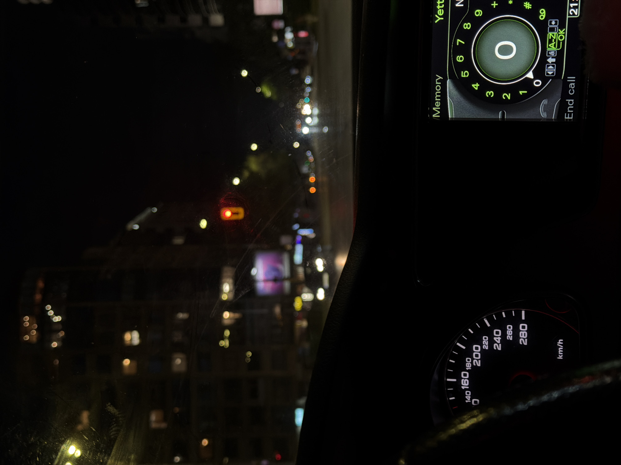 The image presents a nighttime view from the interior of a car, looking out through a windshield that shows some smudges and scratches. The scene outside is a blurred urban landscape illuminated by various artificial lights, indicative of a city at night.
Inside the car, a portion of the steering wheel is visible in the lower right, along with the car's instrument cluster. A speedometer is clearly displayed, calibrated up to 280 km/h, with the needle resting at "0", signifying the vehicle is stationary.
To the upper right, an in-car display screen is active, showing a phone dialer interface. The screen prominently features a large "0" surrounded by a circular numeric keypad (0-9). Visible text on this display includes "Yett", "N", "Memory", "A-Z", "OK", "End call", and "21:", suggesting a time of day around 9 PM.
Outside the vehicle, the dark environment is punctuated by numerous out-of-focus light sources. A distinct red traffic light is visible on the left, indicating the car is stopped. Beyond this, blurred lights from buildings and other vehicles create a bokeh effect. Multi-story buildings with illuminated windows are discernible in the distant background. The presence of "km/h" on the speedometer confirms the car is in a metric-system country, consistent with the provided context of Sofia, Bulgaria. The overall ambiance is that of a quiet, late evening or night in an urban setting.