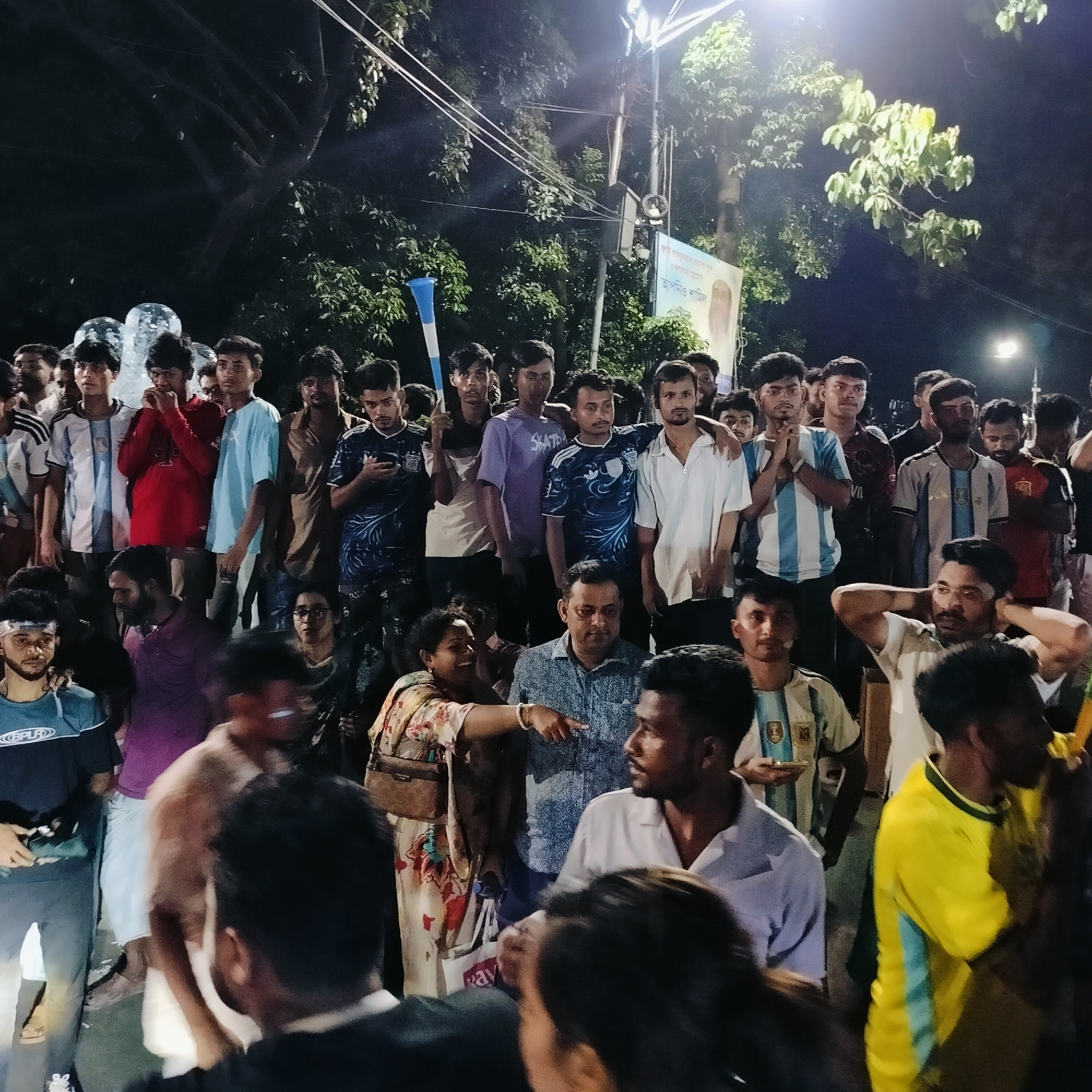 This is a medium shot of a crowd of people, predominantly young men, gathered outdoors at night. Many are wearing football jerseys, suggesting a shared interest or event, possibly related to a match. Several individuals are looking in the same direction, as if watching something or someone. A banner with text and a portrait is visible in the background, with a street lamp illuminating the area. The scene appears to be taking place in a public space in Dhaka, Bangladesh, given the presence of Bengali script on the banner and the overall context. The lighting suggests it is nighttime, and the dense gathering of people indicates a public event or gathering.