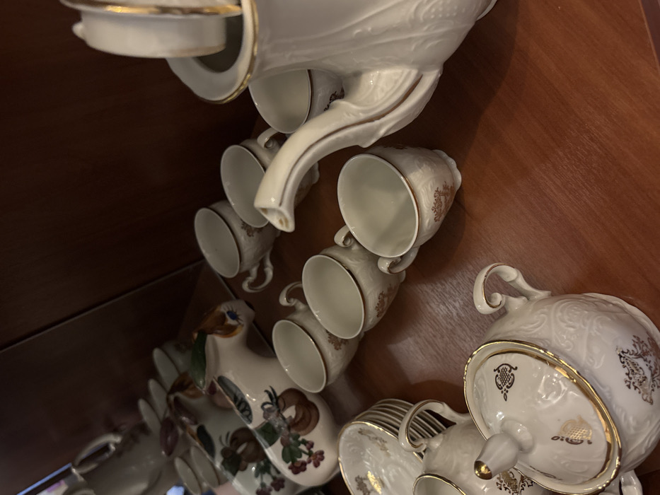 The scene is located in Velikiye Luki, Russia. It depicts a collection of ceramic tableware arranged on a polished, reddish-brown wooden surface within an indoor setting.
On the right side of the frame, a white porcelain coffeepot or teapot with a gold-trimmed lid and spout is prominently featured. To its left and in front, six matching small cups, also white, are visible, each adorned with a gold rim and intricate embossed or painted patterns. In the lower left quadrant, a lidded sugar bowl and a smaller creamer, both white with gold details and decorative designs, are present, along with a stack of matching saucers.
Towards the upper left background, at least two distinct, elongated, animal-shaped ceramic pitchers are arranged. These vessels are white with hand-painted floral motifs and exhibit brown and green accents, including a visible painted eye on one.
No individuals are present, and no actions or interactions are depicted; the objects are static under ambient indoor illumination, casting soft shadows on the wooden surface.