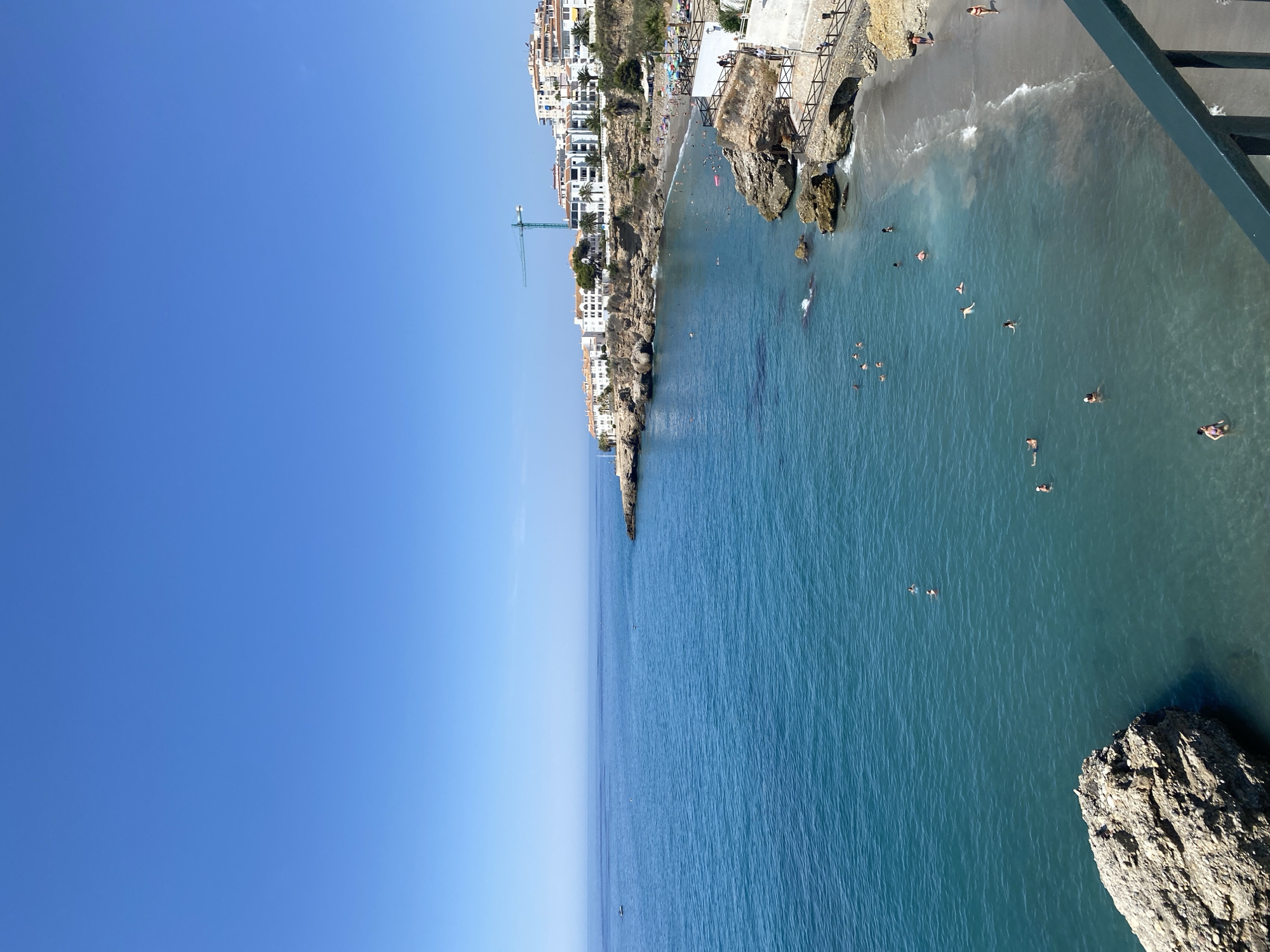 This is a high-angle, full-shot photograph of a beach and coastline in Nerja, Spain. The sky is a clear, vibrant blue with no clouds. The turquoise sea stretches out to the horizon, with gentle ripples on the surface. Several people are visible swimming and wading in the water, with some further out and others closer to the shore.
The coastline is rocky and irregular, with cliffs rising from the sea. A small, sandy beach is nestled between these cliffs, populated by several sunbathers and individuals strolling along the shore. White buildings with terracotta roofs are built along the clifftops, forming a picturesque coastal town. A green construction crane is visible among the buildings.
Various rocky outcrops and formations are present in the water and along the shore. Some of these rocks are large and imposing, while others are smaller. Wooden railings and pathways are visible along the cliff edges, providing access to different vantage points. A dark railing is in the foreground, partially obscuring the view. The overall scene depicts a sunny, active day at a Mediterranean beach.