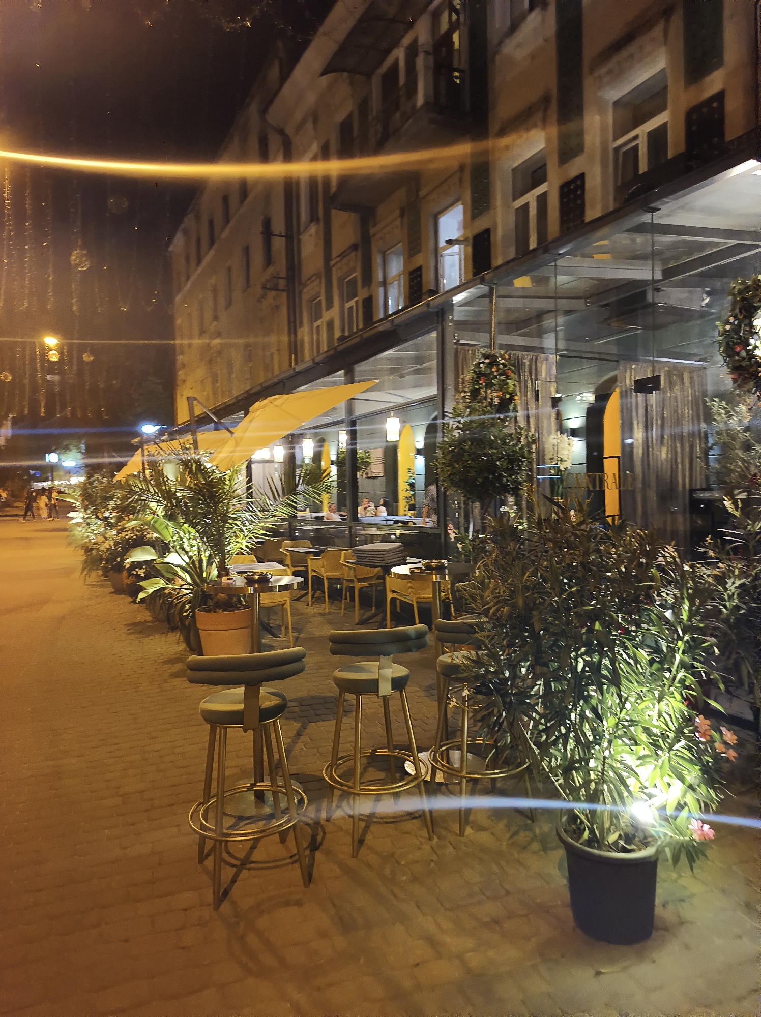 The image depicts an outdoor seating area of a restaurant or cafe at night in Ternopil, Ukraine. The scene is illuminated by artificial lighting, casting warm yellow and white tones on the surroundings. In the foreground, several high bar stools with gold legs and dark green cushions are arranged along a paved walkway. Adjacent to the stools, potted plants, including a large palm-like plant and a smaller plant with pink flowers, are strategically placed and lit.
The seating area is covered by yellow awnings that extend from a building. The building itself appears to be multi-story with windows and architectural detailing. To the right, the establishment features a glass-enclosed section, suggesting an indoor dining area. Inside the glass enclosure, tables and chairs are visible, and some patrons can be seen seated. Draped curtains add a touch of elegance to the interior.
Further down the walkway, more tables and chairs are set up under the awnings. In the background, on the left side of the image, several people are walking along the sidewalk, silhouetted against the ambient light. Streetlights and building lights contribute to the overall illumination of the urban setting. The overall atmosphere is one of a lively evening at a public establishment.