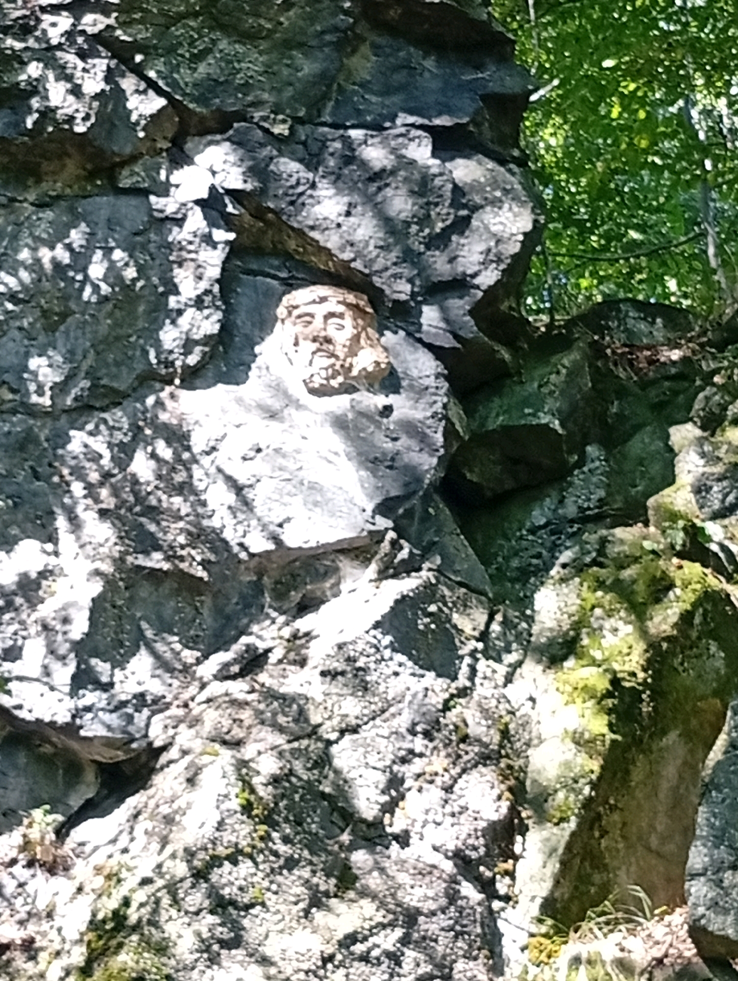 A weathered stone relief of a bearded human face is embedded in a rough, dark grey rock face, positioned at an upward angle. The face, carved with a crown of leaves or vines around its head, exhibits details such as eyes, a nose, and a beard. It is situated in a natural outdoor environment characterized by rugged rock formations and lush green foliage, suggesting a wooded or mountainous setting. The lighting indicates it is daytime, with sunlight casting shadows on the textured surfaces of the rocks. The relief is located on the Góra Ślęża mountain in Sobótka, Poland, a site known for its ancient sculptures and natural beauty.