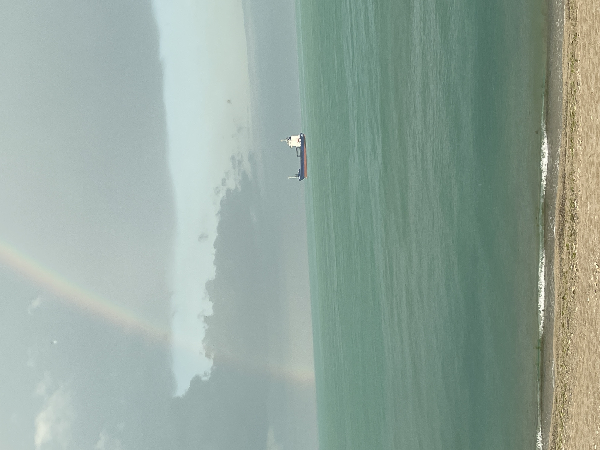The image depicts a view of the Black Sea, likely near Varna, Bulgaria, given the coastal environment and presence of maritime traffic. A cargo ship is visible offshore. A faint rainbow arc is present in the sky. A stretch of sandy beach lines the right edge of the frame.