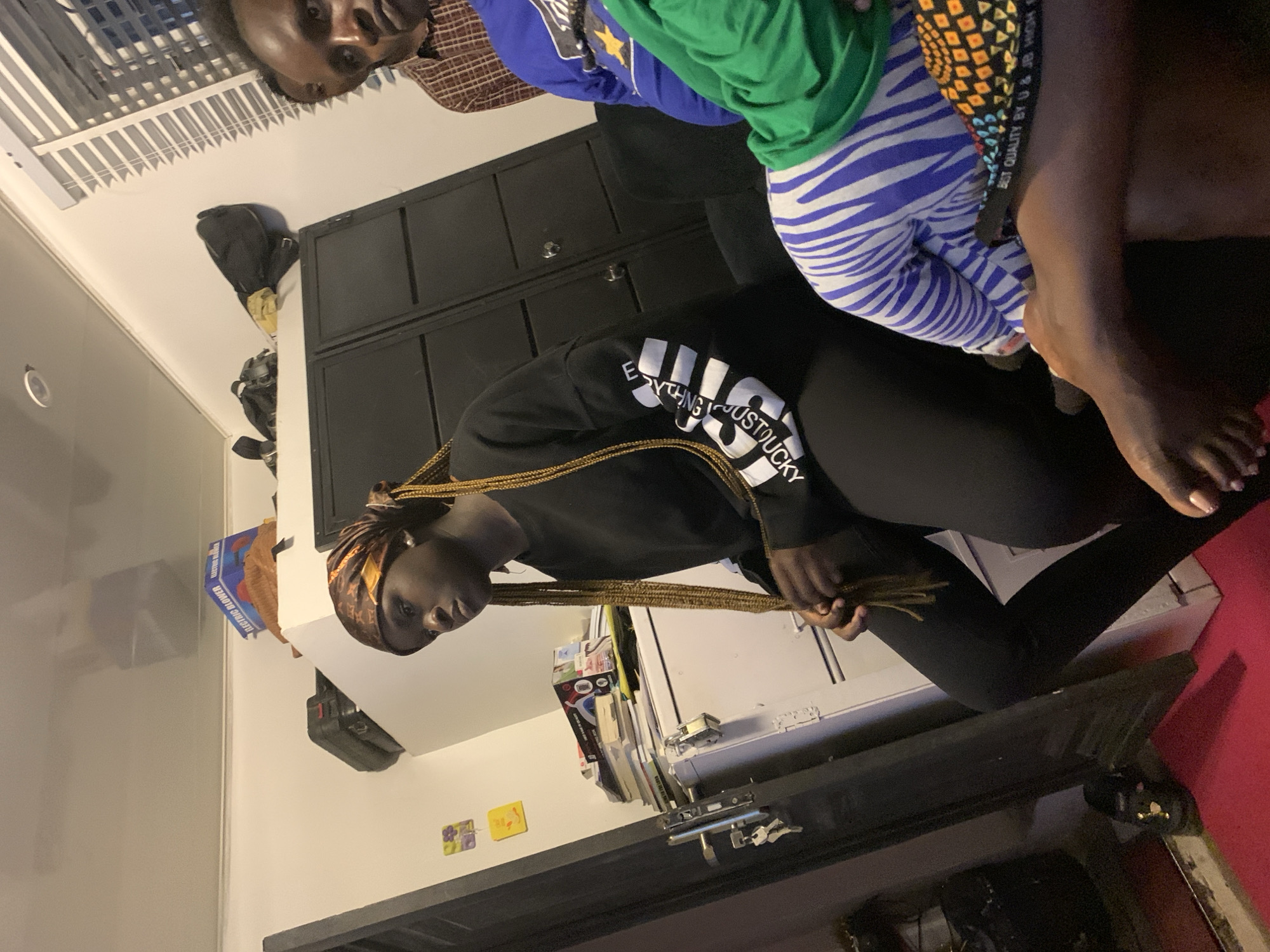 The image depicts an indoor scene featuring four individuals. A young woman in the foreground is wearing a black sweatshirt with white text on the sleeves and a headscarf. She is seated with her legs crossed, looking to her left. To her right, a person in a blue sweatshirt is visible, and next to them, another individual is wearing a green shirt and patterned leggings. In the background, a man in a plaid shirt is partially visible. The environment appears to be a living space or common area, with furniture including a dark cabinet and a white cabinet with books stacked on top. A closed door with a lock and keys is present on the left side of the image. The lighting suggests an indoor setting, possibly during the day or evening. There are no discernible elements that place the scene in a specific city.