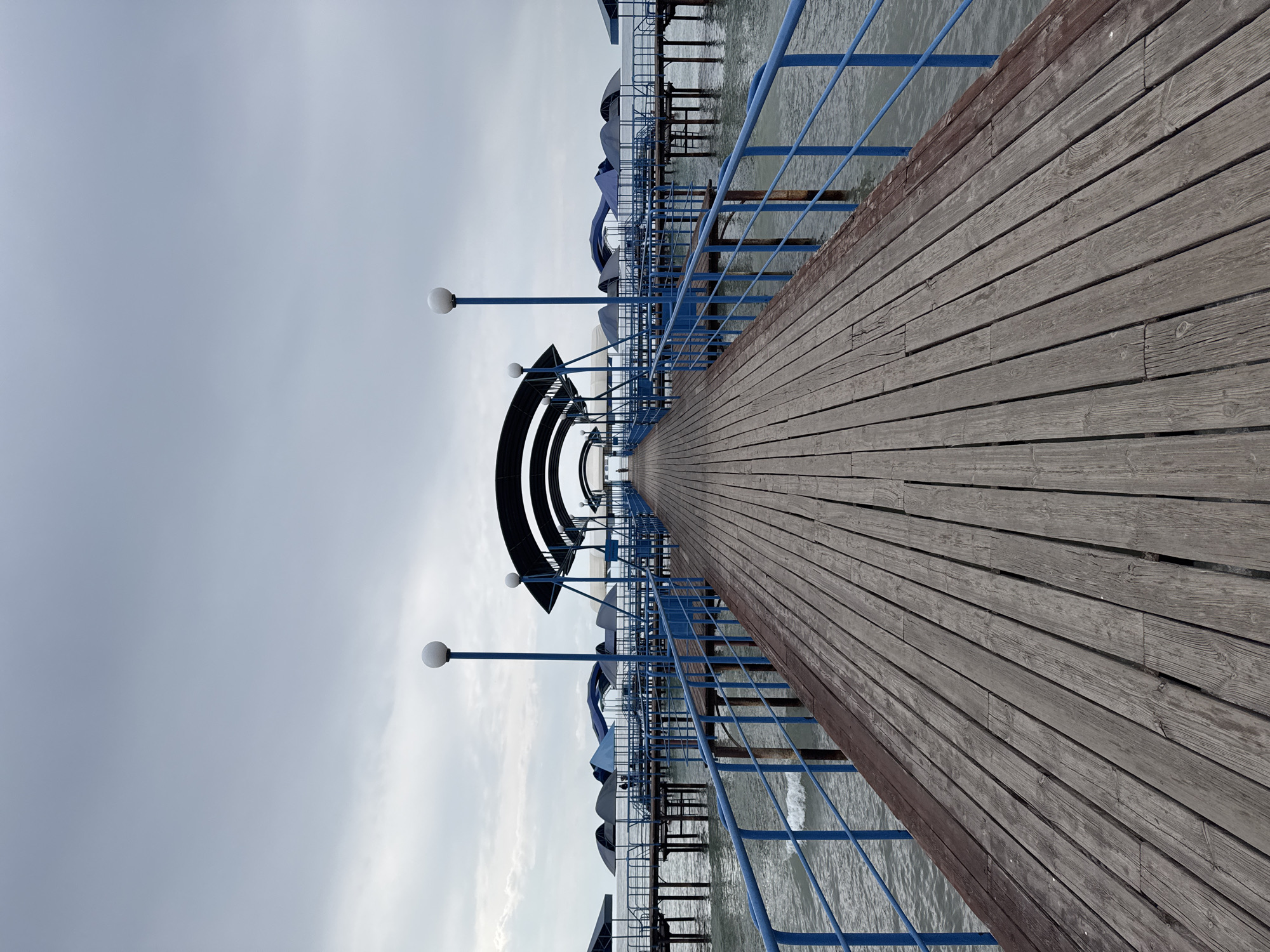 The image shows a long, weathered wooden pier extending out over the water in Sary-Oy, Kyrgyzstan. The pier is lined with blue railings and occasional lamp posts topped with white globes. Arching blue structures with dark roofs are visible along the sides of the pier, creating covered areas. The water beneath the pier is a muted blue-gray, with small waves visible. The sky is overcast with heavy, gray clouds. In the distance, at the far end of the pier, a few indistinct figures can be seen. The overall atmosphere is somber and quiet, typical of a cloudy day.