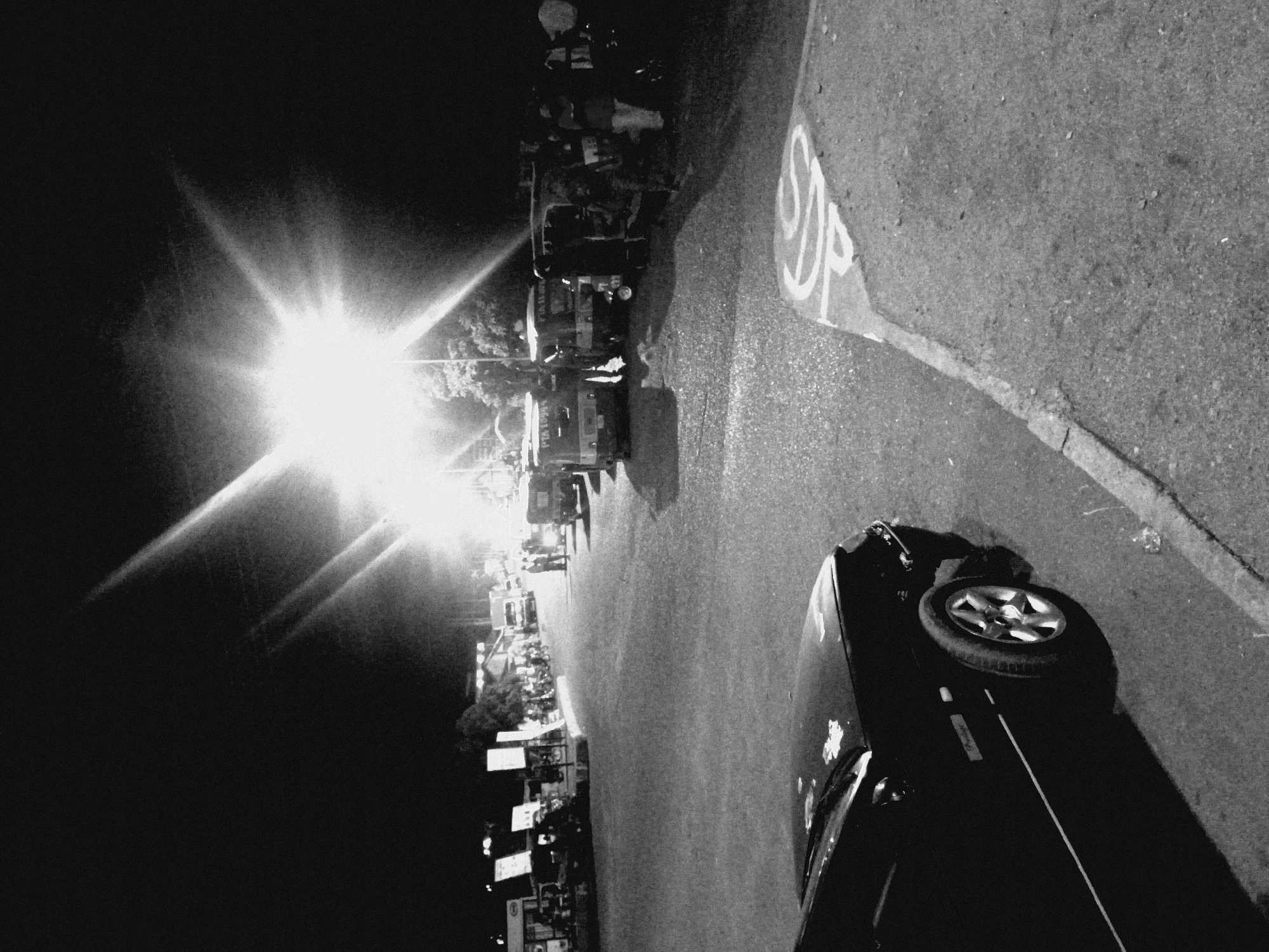 The image shows a street scene in Bukuru, Nigeria, at night. A bright light source dominates the left side of the frame, creating a lens flare effect. The road is visible, with a "SDP" marking painted on the asphalt. Vehicles, including vans and cars, are parked along the roadside. People are present near the vans, though their details are obscured by the darkness and the light. A portion of a car is in the lower right corner. Signs and electrical poles are visible along the roadside. The scene appears to be active, potentially a market or gathering place.