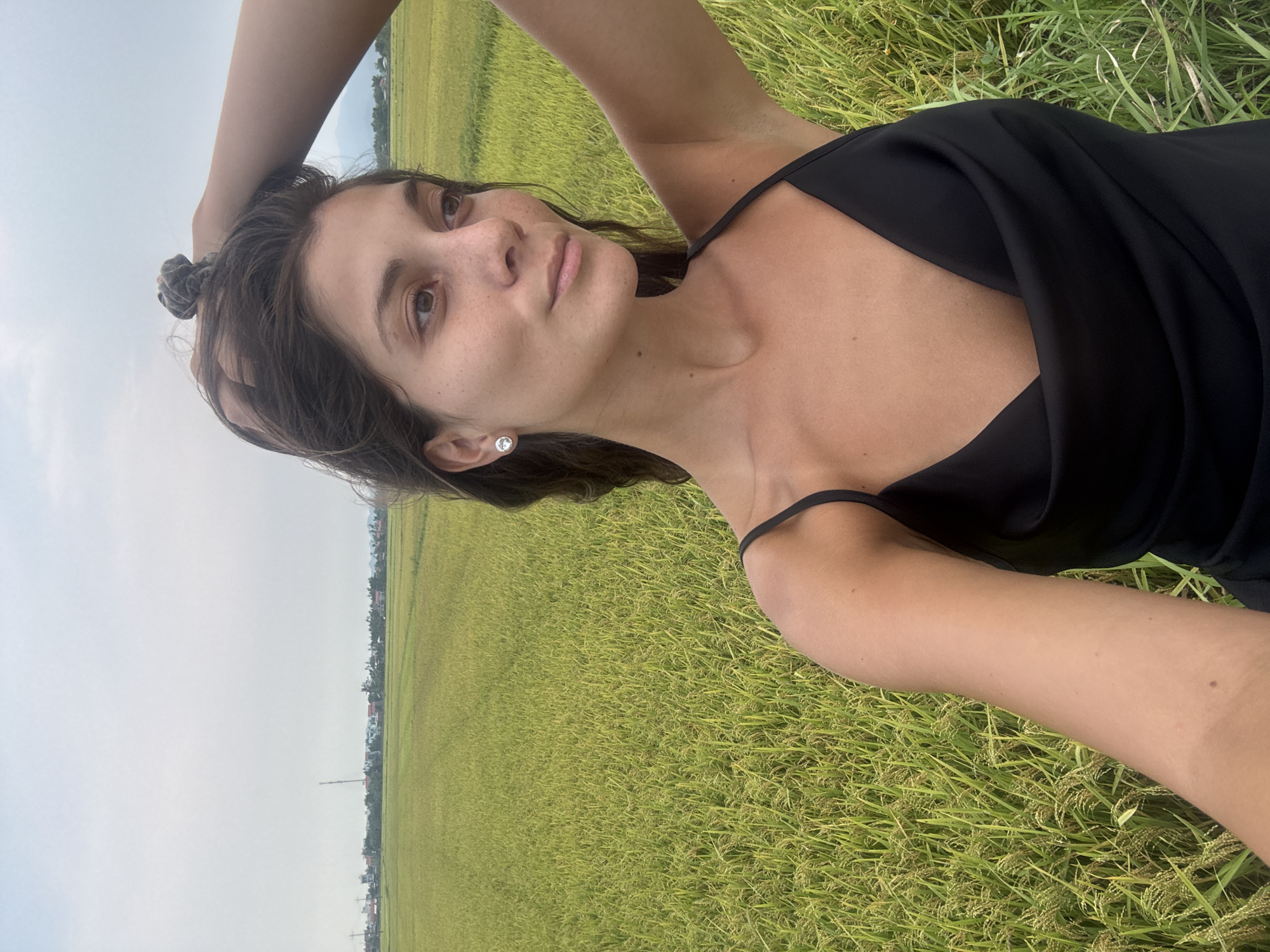 A young woman is prominently featured in this outdoor portrait, positioned as if lying on her back in a vast green field and looking directly upwards towards the viewer. She has medium-brown hair, which appears to be tied up with a scrunchie, and wears a small, sparkly stud earring in her visible left ear. Her complexion is natural with visible freckles, and she has dark eyes and soft facial features. She is wearing a sleeveless black garment, possibly a dress or top, with thin straps visible on her shoulders. Her right arm is bent, with her hand resting on or near the back of her head.
The activity captured is a posed photograph, with the subject maintaining a calm and relaxed expression.
The setting is a large agricultural field, strongly suggestive of a rice paddy due to the specific appearance of the green plants with visible grains, consistent with the location context of Đà Nẵng, Vietnam. The field stretches into the distance under a light, overcast sky. On the horizon, faint outlines of buildings, possibly houses or small structures, and a tall antenna or tower are visible, indicating a blend of rural and possibly semi-urban landscape. The lighting suggests daytime, likely late morning or afternoon, with diffused light under the cloudy sky. There is no visible text in the image.