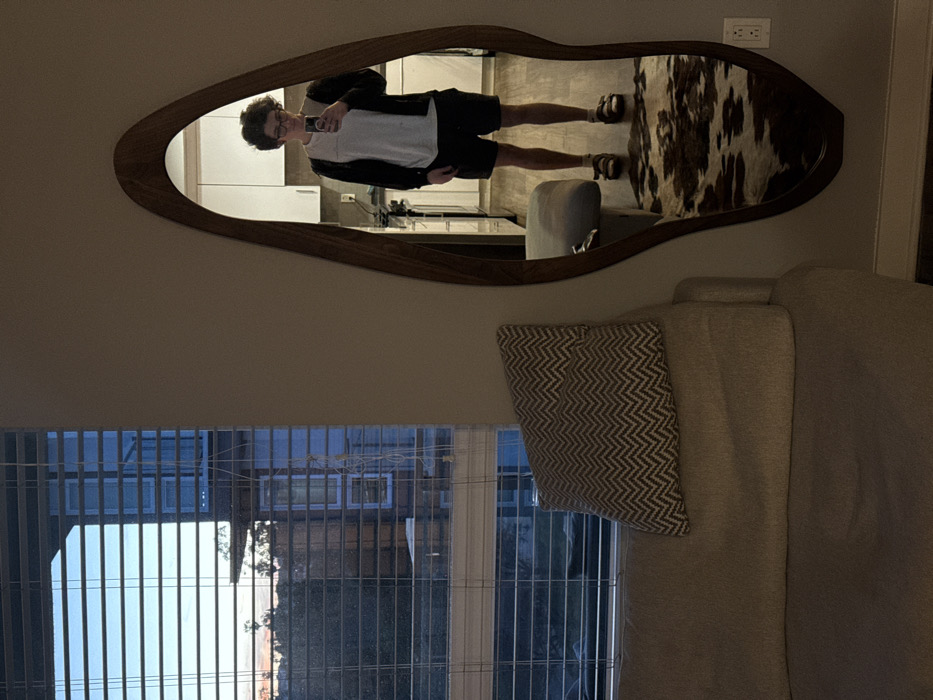 The image displays an indoor scene within a modern residential unit, characteristic of an urban environment in Los Angeles, United States of America. A young adult, with curly dark hair and glasses, is visible taking a selfie in a large, irregularly shaped mirror with a dark wood frame. The individual is wearing a white short-sleeved shirt, a dark long-sleeved open cardigan, dark shorts, and dark sandals, holding a smartphone in their hands at chest level.
The reflection in the mirror reveals a contemporary kitchen area behind the person, featuring white upper cabinets and light-colored countertops. The floor visible in the reflection is a wood-look material, and a patterned rug, resembling cowhide with brown and white patches, is placed on the floor directly beneath the mirror. A white electrical outlet is mounted on the wall to the right of the mirror.
On the left side of the image, a segment of a light-colored upholstered sofa is visible, topped with a throw pillow featuring a white and light brown/grey chevron pattern. A large window, fitted with horizontal white Venetian blinds, occupies the upper left portion of the frame. Through the partially open blinds, an urban landscape is discernible, showing the facades of other multi-story residential buildings, some with visible windows and balconies, along with trees and foliage. The sky outside exhibits the gradient colors of late afternoon or early evening, transitioning from blue to a soft orange and pink at the horizon. The interior lighting is subdued, suggesting natural light is fading.
