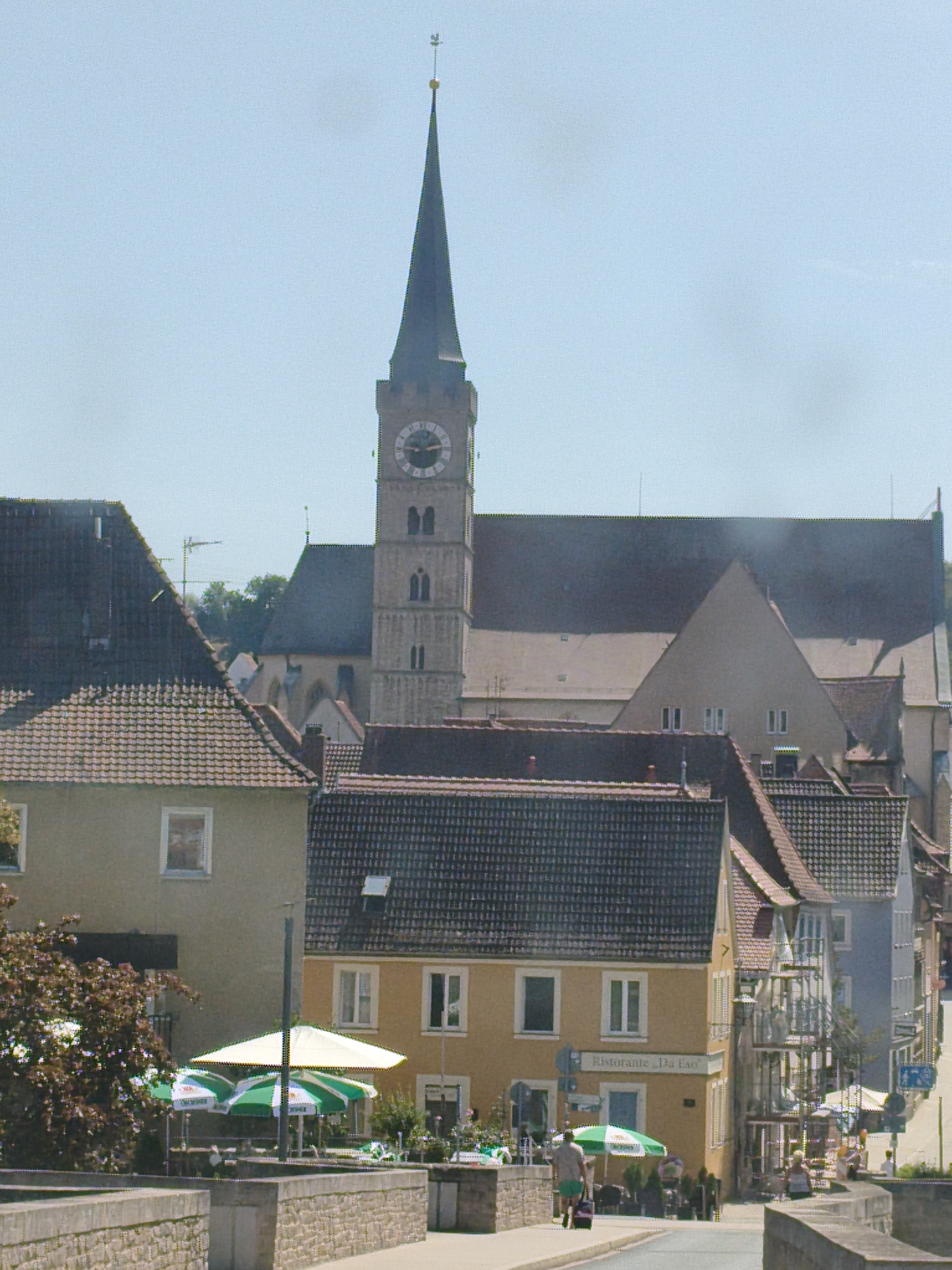 This is an eye-level, full shot of a European town on a clear day. In the background, a tall, Gothic-style church tower with a clock face dominates the skyline. Below the tower, various buildings with tiled roofs and light-colored facades line a street. In the foreground, a stone wall runs across the bottom of the frame. To the left of the wall, a small tree with dark green leaves is visible. In the middle ground, a yellow building with awnings and outdoor seating suggests a cafe or restaurant. Several people are scattered throughout the scene, some walking, some seated at tables. The overall impression is one of a peaceful, urban environment. The image appears to be of Ochsenfurt, Germany, a town known for its historic architecture and scenic location on the Main River.