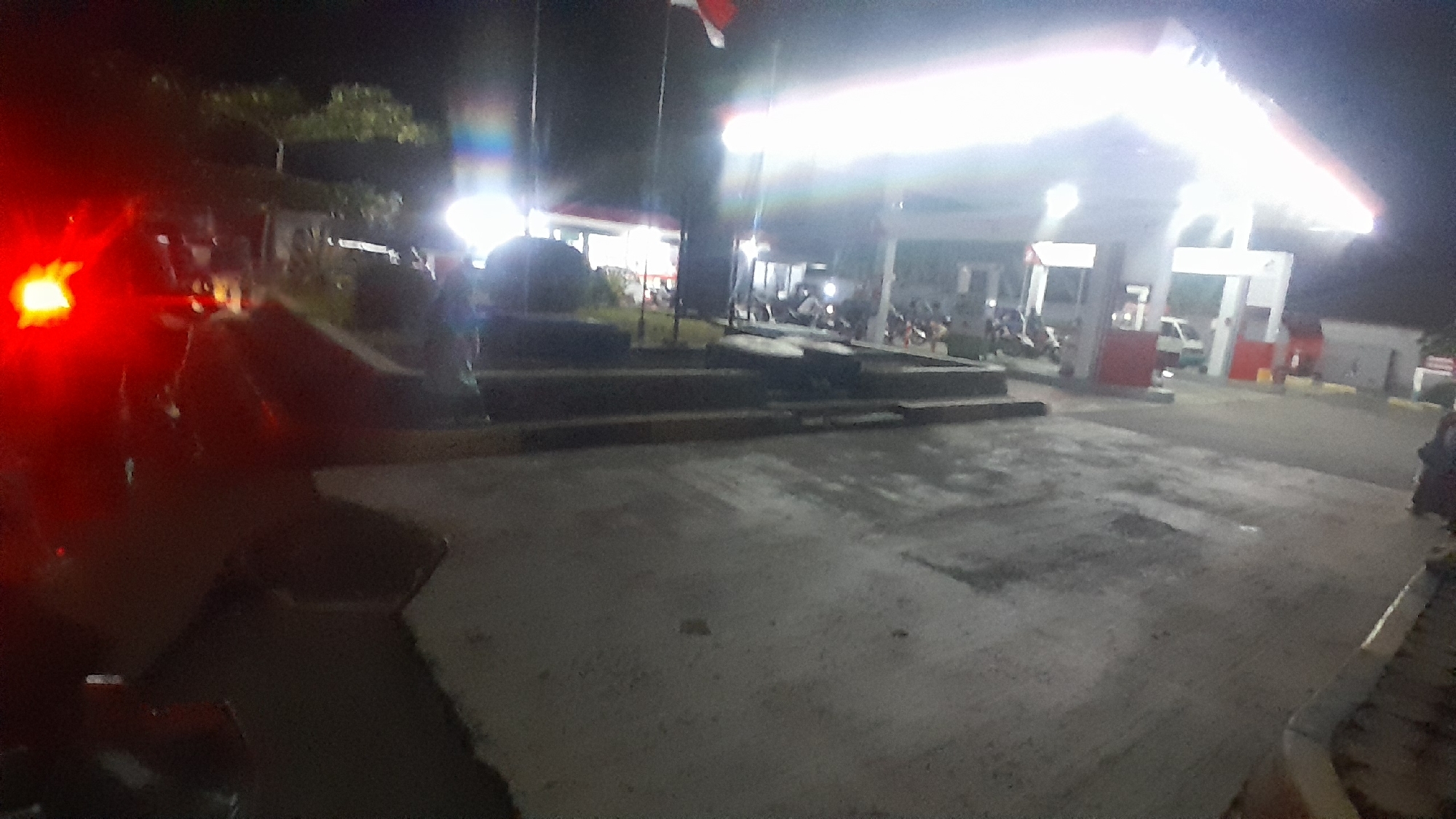 The image depicts a nighttime scene at a gas station in Subang, Indonesia. The gas station is well-lit, with bright fluorescent lights illuminating the canopy and the surrounding area. Several vehicles, including motorcycles and a small van, are visible at the pumps. People appear to be present, likely customers and employees, though their specific activities are obscured by the low light and image quality. In the foreground, a portion of a red vehicle's taillight is brightly illuminated, suggesting it is positioned close to the camera and potentially in motion. The ground is a mixture of concrete and asphalt, with some areas appearing wet or stained. Trees and vegetation are visible in the background, silhouetted against the night sky. A flag, likely the Indonesian flag, is visible flying from a pole. The overall impression is one of a busy, functional gas station operating during evening hours.