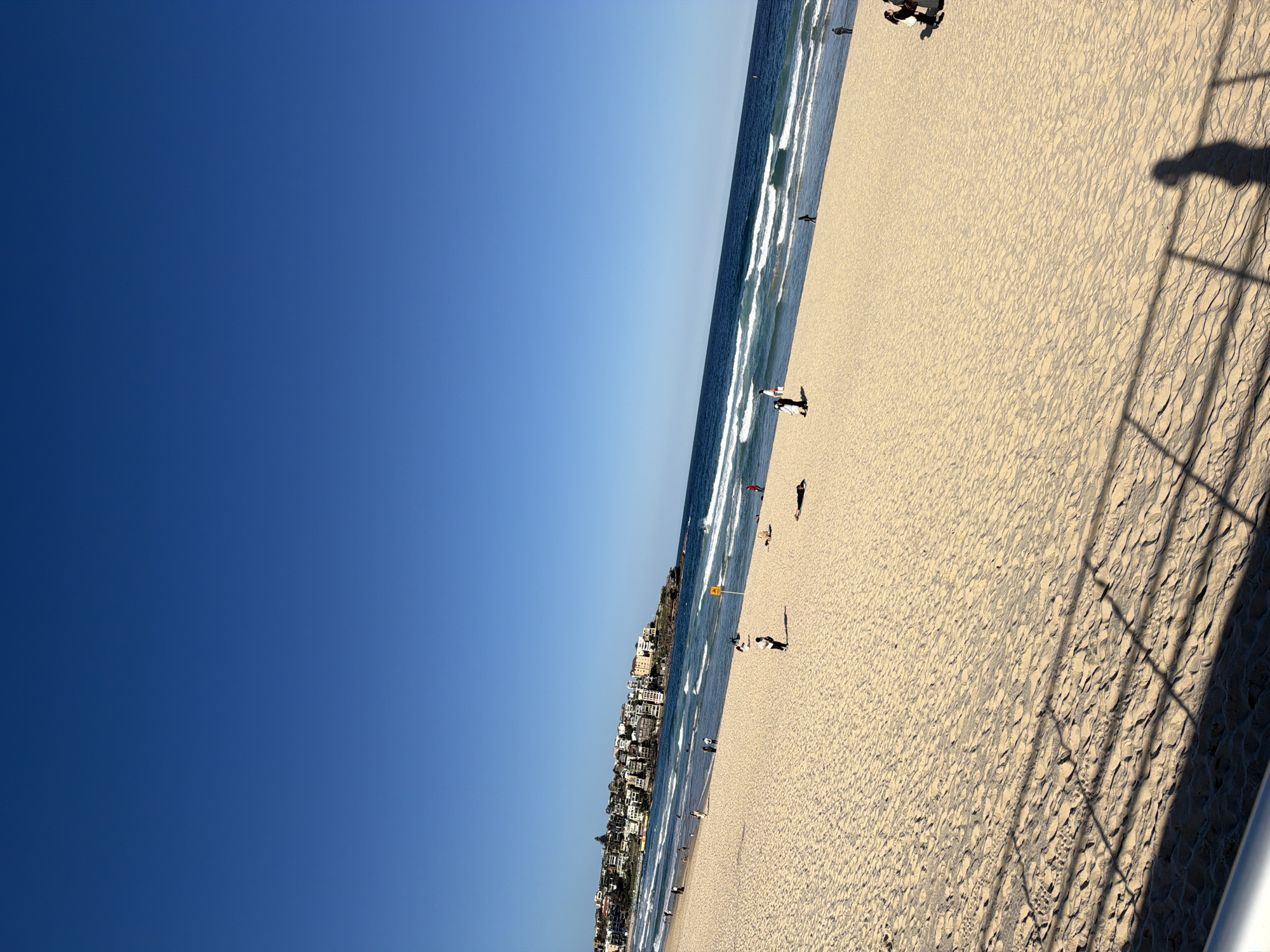 The image depicts a wide, sandy beach at Bondi Beach, Australia, under a clear, bright blue sky. In the distance, the characteristic buildings of the Bondi coastline are visible, curving along the shore. Gentle waves roll onto the beach from the deep blue ocean. Several individuals are scattered across the sand, with some walking near the water's edge, while others are reclining or standing further up the beach. A yellow flag is visible in the distance, likely indicating a swimming zone. The foreground shows the textured sand and the shadow of a railing cast across it. The overall scene conveys a peaceful, sunny day at a popular beach location.