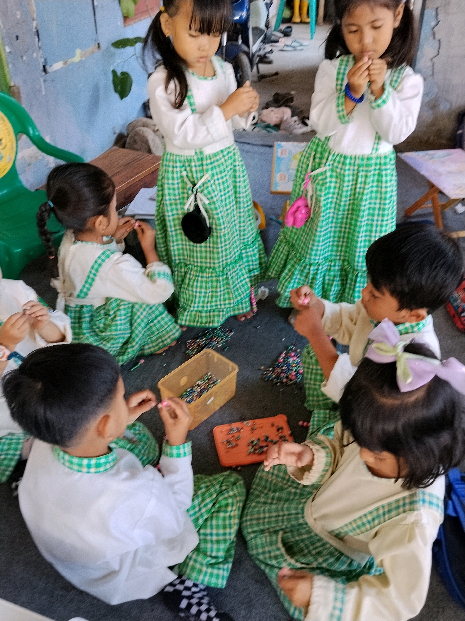 In Panggungan, Indonesia, a group of young children, likely students, are seated on a dark grey floor, engaged in an activity with small, colorful beads. They are all wearing matching outfits consisting of white long-sleeved tops with green plaid trim and matching green plaid skirts or overalls.
In the foreground, a young boy with black hair is reaching for a pink bead, his attention focused on the task. To his right, a young girl with dark hair adorned with a light purple bow is also reaching for beads.
Behind them, two other children are seated. One, a boy with dark hair, is holding a pink bead in his hand, looking down. Another child, mostly obscured, is wearing the same uniform.
In the background, two girls are standing. The girl on the left, with dark hair in pigtails, is looking down, possibly at beads in her hands. The girl on the right, also with dark hair, is holding beads near her mouth.
The environment appears to be a simple room with a concrete floor, a blue-painted wall in the background, and some wooden furniture. There is a green plastic chair on the left, a wooden table with items on it in the middle background, and another small wooden table on the right. Several containers hold beads, including a yellow rectangular bin and an orange rectangular tray. Various loose beads are scattered on the floor around the children. The overall scene suggests an educational or craft activity taking place.