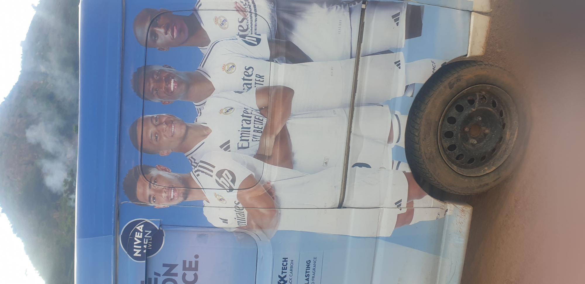 The image depicts the side of a blue van, serving as a mobile advertisement. The prominent graphic on the van features four young men in white Real Madrid football jerseys. The man on the far left is smiling and looking to the right, his arms crossed. Next to him, a man with short dark hair is also smiling and looking forward. The third man from the left, with dark skin and short hair, has a wide smile and is looking forward. The man on the far right, with dark skin and closely cropped hair, is looking forward. Above the players, a NIVEA MEN logo is visible. The lower portion of the van shows part of a wheel and tire. The background consists of a hazy, mountainous landscape, possibly indicating an outdoor setting. The van appears to be parked on a dirt surface, with dust kicked up around the wheel. The text on the van includes "MIRATES," "FLY BETTE," and other unreadable fragments. The overall impression is a promotional vehicle, likely circulating within a city or town. Given the context, this image was captured in the city of Jos, Nigeria.