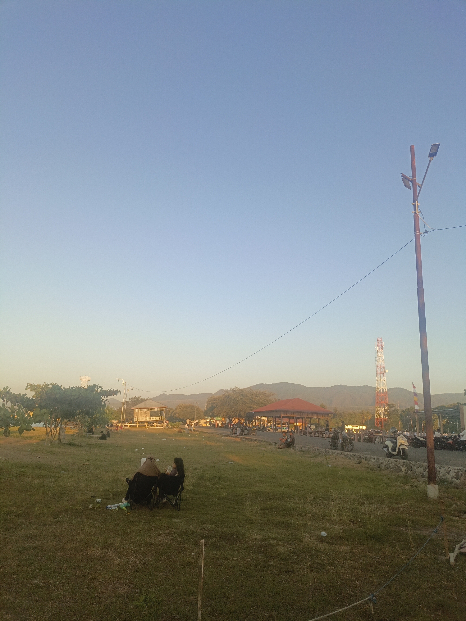 This image depicts an outdoor scene in the city of Lewoleba, Indonesia, likely during the late afternoon or early evening due to the warm, soft lighting. In the foreground, a grassy field occupies the majority of the frame. Two individuals are seated in camping chairs, facing away from the camera, appearing to be relaxing and conversing. They are positioned in the lower left quadrant of the image. Scattered on the grass near them are some personal items, including what appears to be a blue and white cooler bag and possibly some snacks.
The middle ground reveals a paved area with several parked motorcycles, suggesting a public gathering or recreational space. A distinctive red-roofed structure, possibly a pavilion or open-air dining area, is visible, with people milling around it. Further in the background, a prominent red and white telecommunications tower stands tall against the sky, indicating the presence of infrastructure. Beyond the tower, rolling hills form a mountainous backdrop. A solitary lamppost is positioned on the right side of the frame, near the edge of the paved area. The sky is clear and a pale blue, transitioning to a lighter hue towards the horizon, suggesting the setting sun. The overall atmosphere is serene and leisurely, with a sense of community engagement in an open, natural setting.