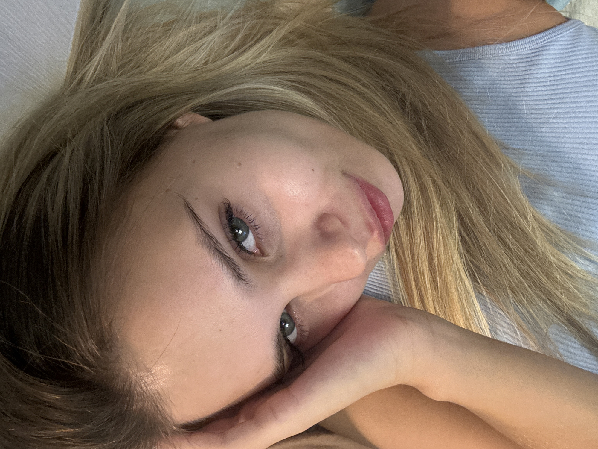 The image features a close-up portrait of a young woman with long, blonde hair and light blue-green eyes. She is lying down, with her head tilted, looking directly towards the viewer. Her left hand is resting near her chin and cheek, supporting her head slightly. She has a serene or contemplative expression.
She is wearing a light blue, ribbed top or shirt, visible around her neck and shoulder. Her blonde hair is spread out, covering parts of her shoulders and the background. The lighting is soft, suggesting an indoor setting, likely during the daytime. The background is out of focus, appearing to be a light-colored surface, possibly a bed or pillow. There is no visible text in the image. The overall impression is one of calm and introspection.