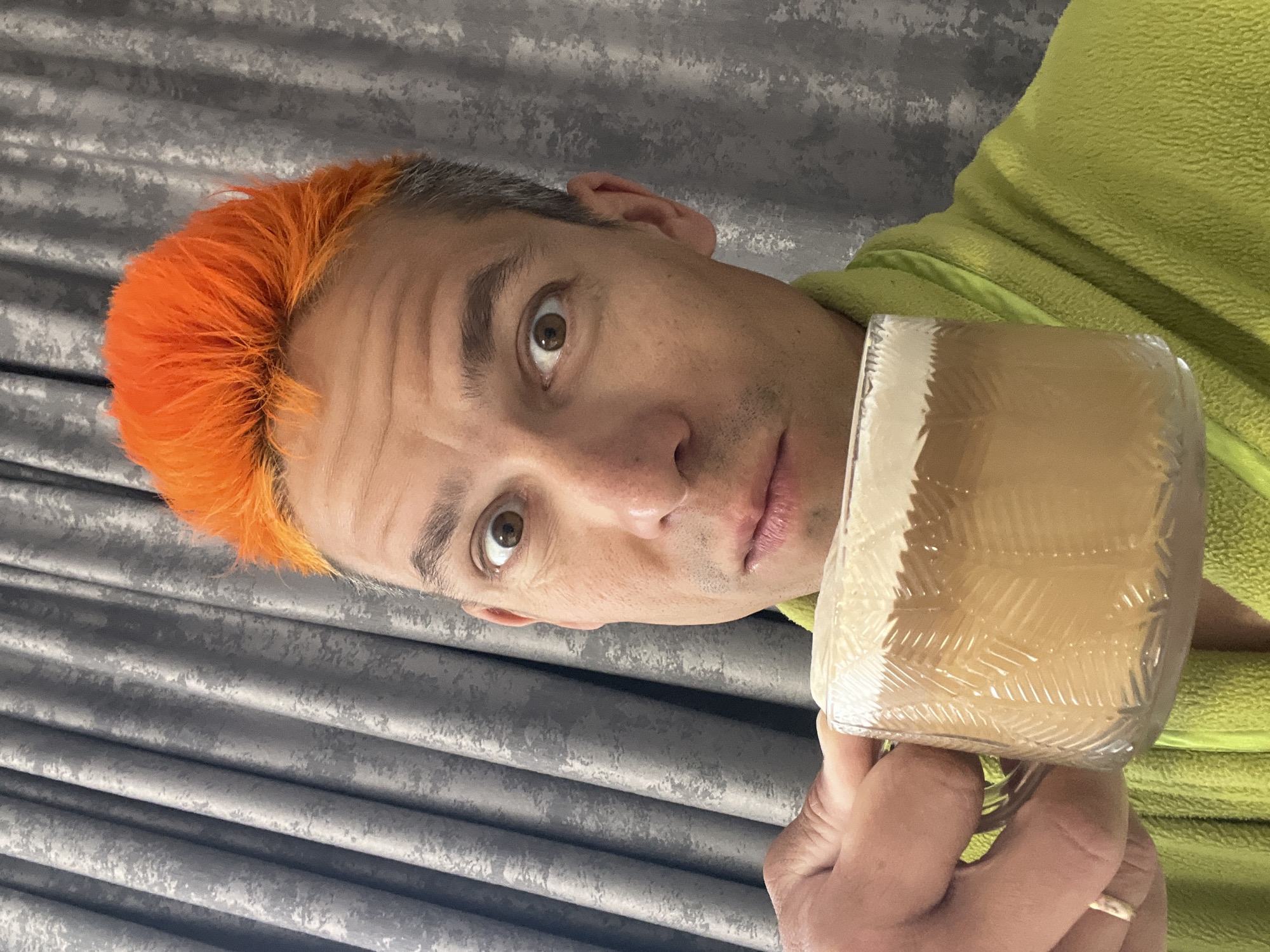 A light-skinned person with orange hair is holding a clear glass in their right hand. The glass appears to be filled with a light brown liquid with a white foamy top. The person wears a light green fleece jacket or robe. Behind the person is a gray textured wall or curtain. The person's expression is neutral, and they are looking directly at the viewer. The photograph does not provide enough information to determine its location in the city of Chaikovsky, Russia.