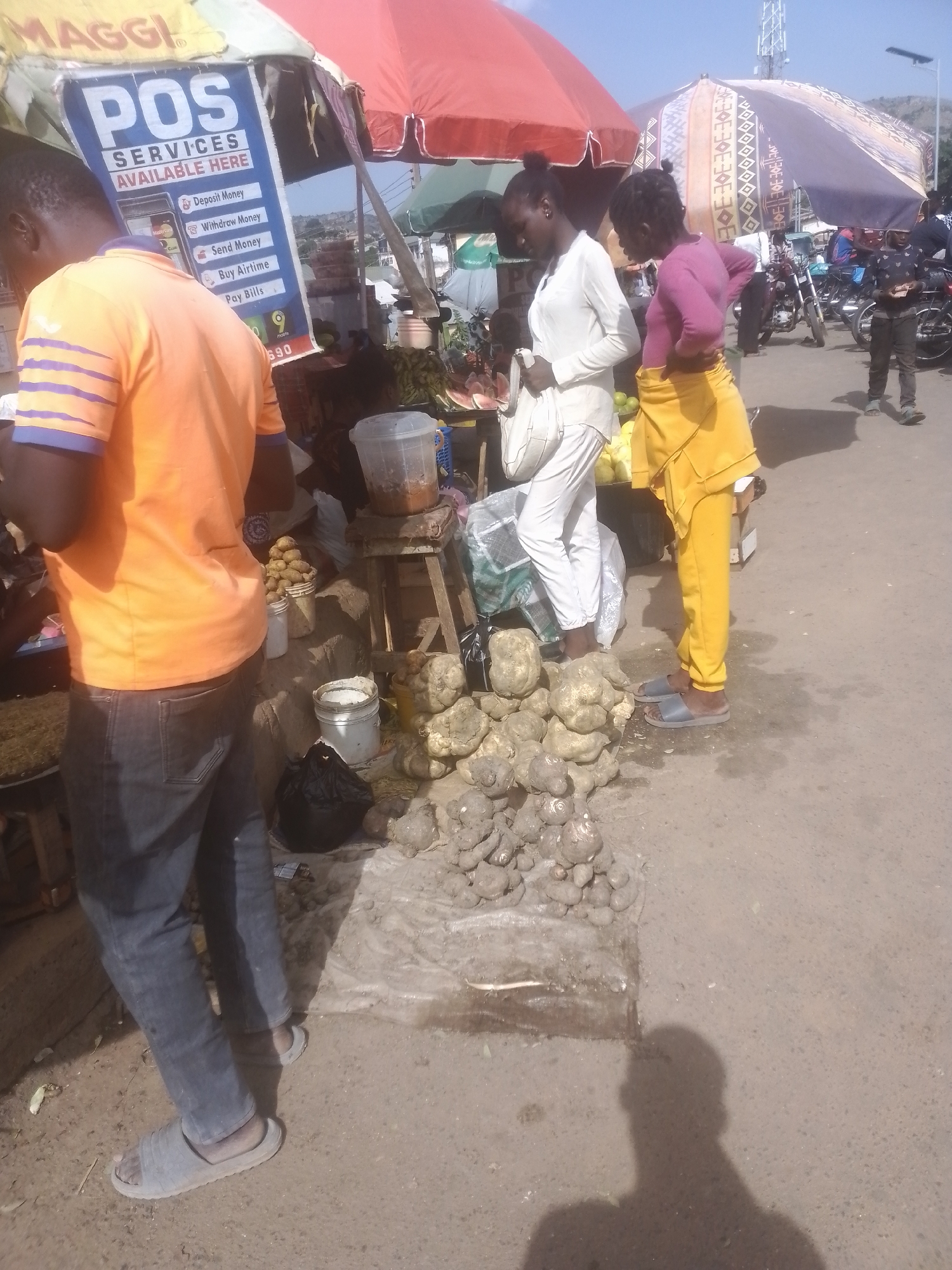 This media file captures a vibrant outdoor market scene, likely in a tropical or subtropical climate given the bright sunlight and clear sky. The primary subjects are several individuals engaged in a transaction or conversation at a market stall. One person, wearing an orange shirt and blue jeans, stands with their back to the camera, looking towards the stall. Next to them, a woman in a white top and white pants is looking down, while another woman in a pink top and yellow pants is also looking towards the stall.
The market stall itself is laden with produce, including what appear to be root vegetables like yams or cassava piled on the ground. Several containers, including a large clear plastic one with a dark substance inside and white buckets, are also visible. Behind the stall, a large umbrella provides shade, with a "MAGGI" logo visible on a sign hanging from it. Another sign for "POS SERVICES AVAILABLE HERE" details various services offered, such as "Deposit Money," "Withdraw Money," "Send Money," "Buy Airtime," and "Pay Bills."
In the background, more market stalls, umbrellas, and a road with motorcycles suggest a bustling commercial area. The time of day appears to be daytime, with strong shadows cast by the sun, indicating a bright and possibly warm day. The overall atmosphere seems busy and transactional, typical of a local marketplace. The visible text provides specific details about the services offered at the POS stand, highlighting financial services accessible at this location.