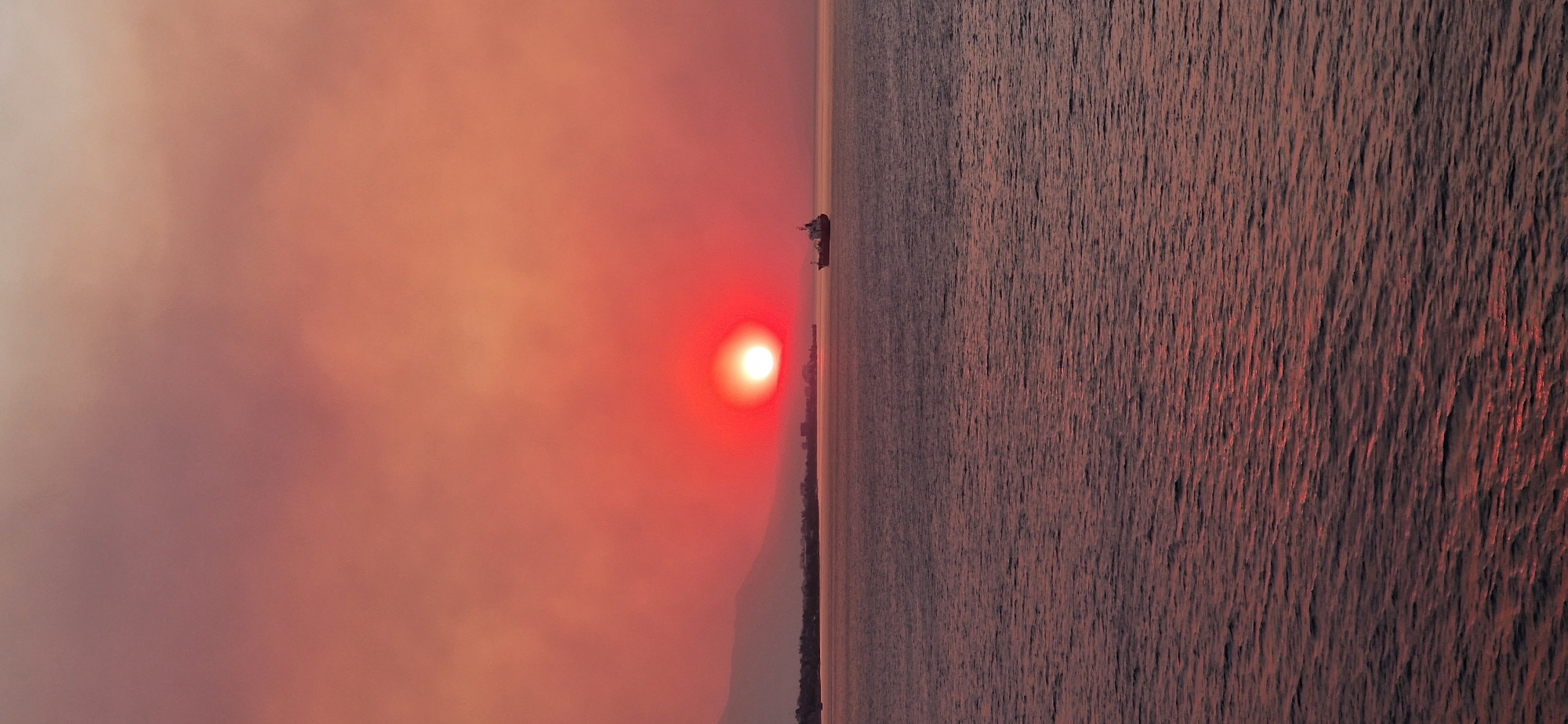 A cargo ship sails on the sea during a dramatic sunset over the Municipal Unit of Agioi Theodoroi, Greece. The sun, a bright orange-red orb, is partially obscured by atmospheric haze, casting a fiery glow across the sky. The water reflects the warm hues of the sunset, creating shimmering ripples. In the distance, the coastline is visible with silhouetted buildings and mountains. The ship, a solitary dark shape against the luminous backdrop, is positioned in the mid-right of the frame. The overall scene is one of serene, natural beauty, with a touch of industrial presence from the ship.