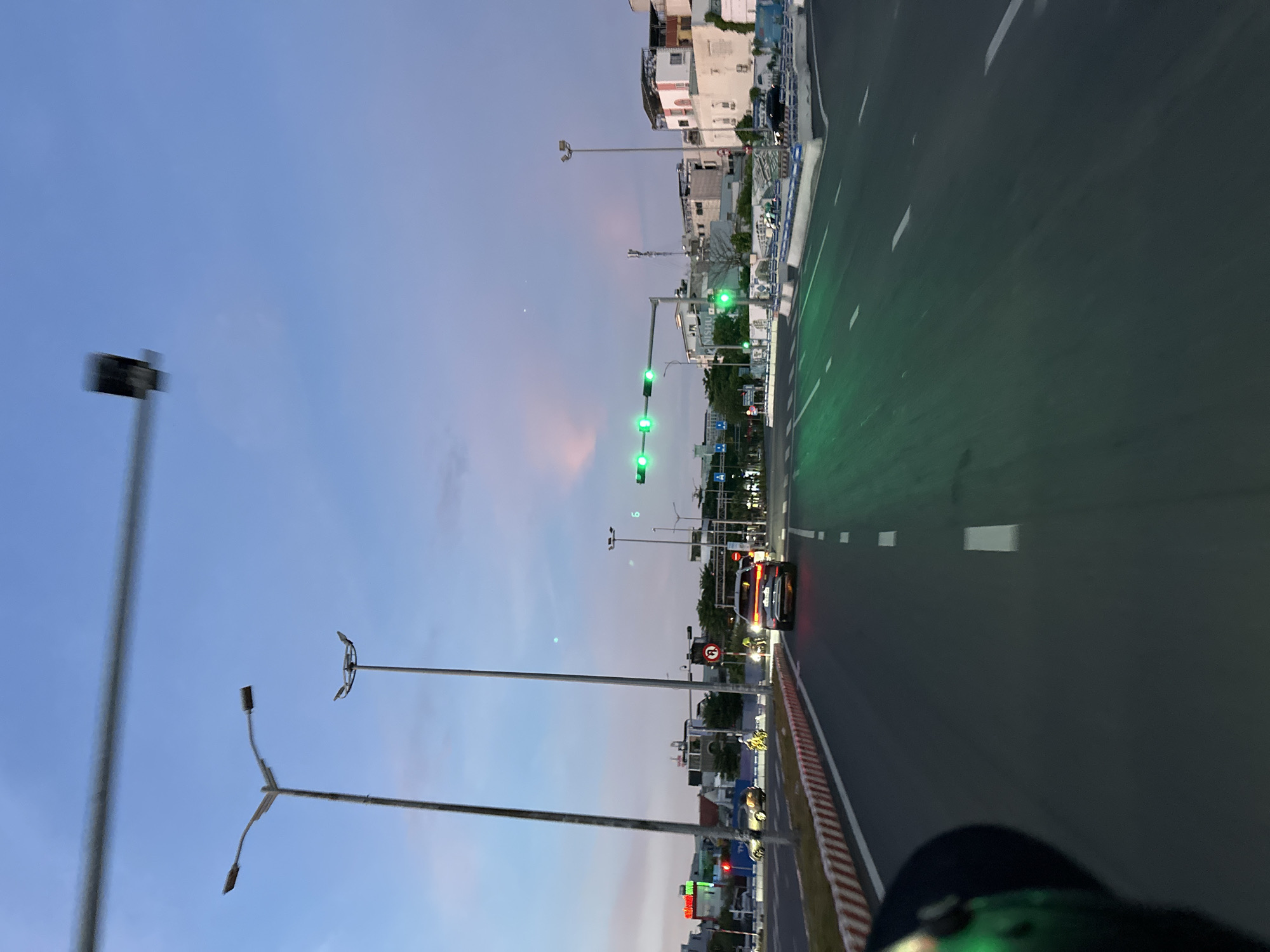 The image captures a street scene in Da Nang, Vietnam, during the twilight hours. The sky displays a gradient of blue and pink hues, suggesting the transition from day to night. On the road, a dark-colored SUV is visible, its taillights illuminated. Several traffic lights are clearly visible, with their green lights illuminated, indicating that traffic is permitted to proceed.
Tall streetlights line the sides of the road, their poles extending upwards into the sky. In the background, buildings of various sizes and architectural styles are present, some appearing to be residential or commercial structures. The road itself is paved and marked with white lane dividers. There is a concrete median with red and white striped barriers separating lanes.
The perspective suggests the image was taken from a moving vehicle, likely a motorcycle or a car, as indicated by the blurred foreground element on the left and the motion blur present throughout the scene. The overall impression is that of a bustling urban environment at dusk, with vehicles and infrastructure typical of a Southeast Asian city.