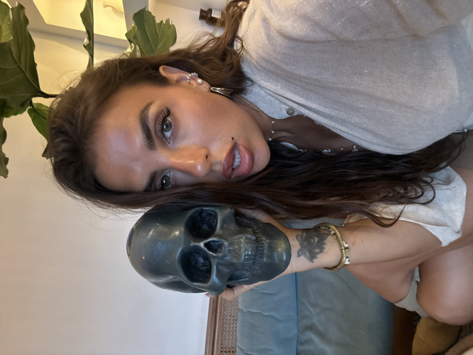 A woman is holding a dark-colored skull in her hands. She has long, dark hair and is wearing a light-colored, textured top. A plant with large, green leaves is visible in the background. The woman is wearing jewelry including earrings and a bracelet. A tattoo is visible on her arm. A blue couch or cushion is present in the background. It is not possible to confirm the location as North Kuta, Indonesia based on visual evidence.
