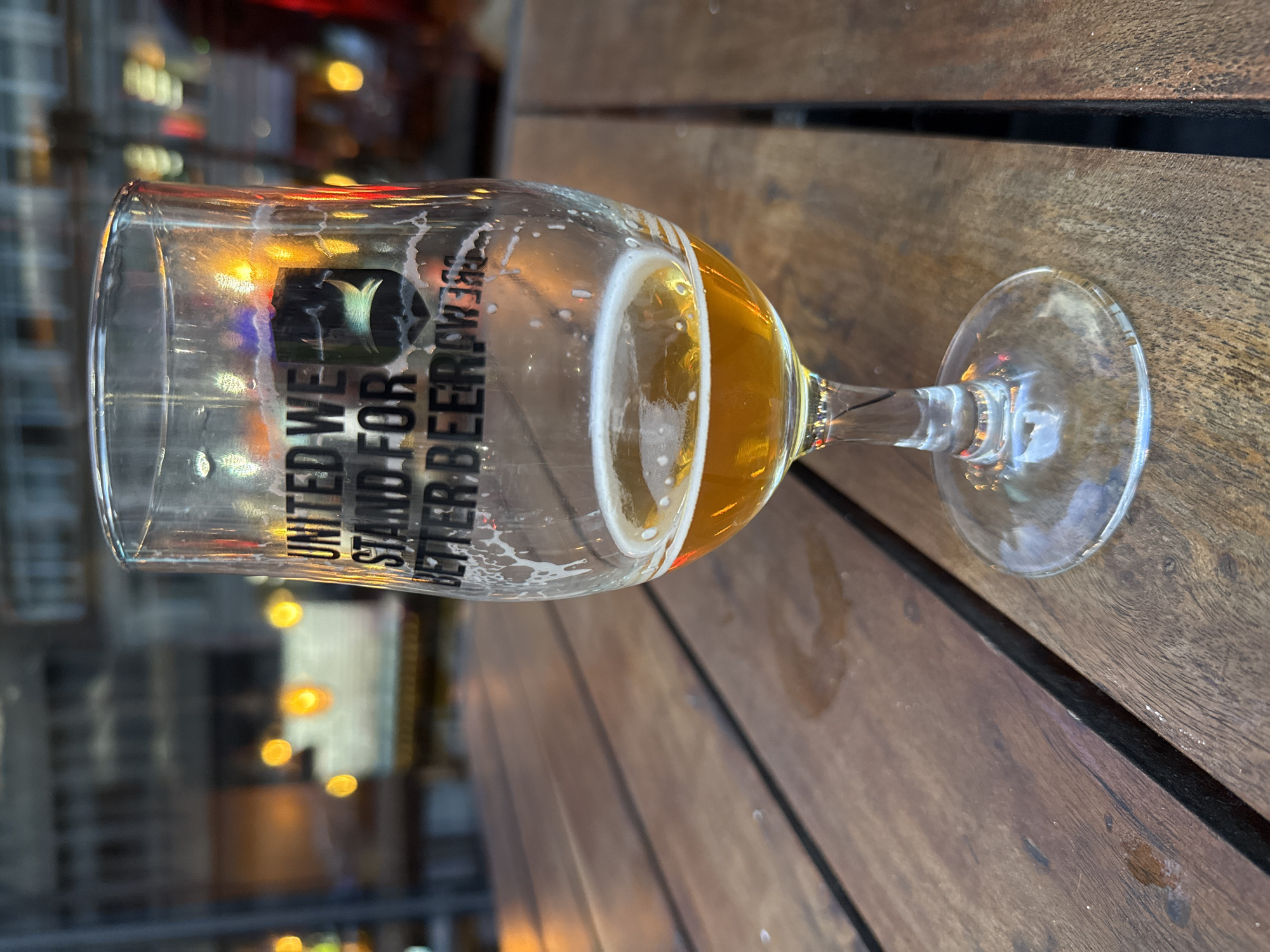 A nearly empty glass of beer rests on a wooden table, presumably at an outdoor pub or restaurant. The glass has the Brewdog logo and the words "UNITED WE STAND FOR BETTER BEER" printed on it. The remaining beer is golden in color, with some foam residue clinging to the glass. A few scattered drops of liquid are on the table near the glass. In the blurry background, lights from buildings suggest an urban setting. Based on the style of the pub and the brewdog logo, the location is likely to be in the city of London, United Kingdom. There are no visible people or distinct actions captured in the photograph. The focus is solely on the beer glass and its immediate surroundings.