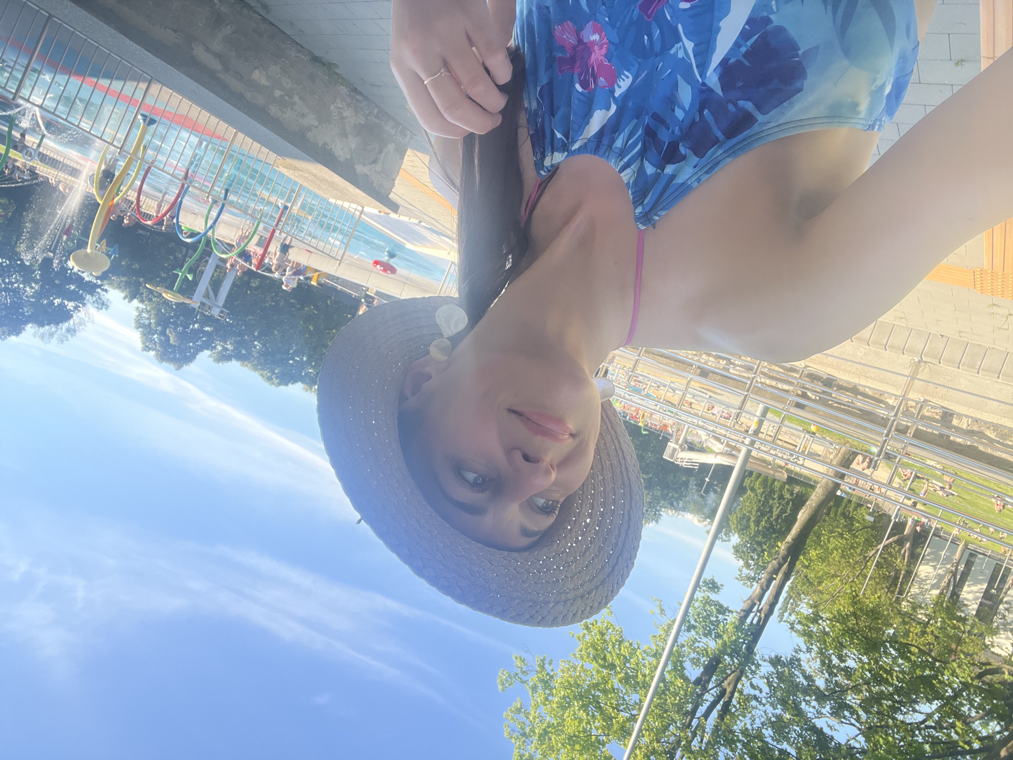 The image is a selfie of a woman wearing a blue floral swimsuit and a straw hat. She is holding her hair back with her left hand, revealing a ring on her finger. The woman is smiling and looking at the camera. The background shows a swimming pool area with colorful water features and people relaxing. The sky is blue with some wispy clouds. The image appears to be taken on a sunny day, likely in the afternoon, given the lighting. The overall mood is cheerful and relaxed, suggesting a vacation or leisure activity.