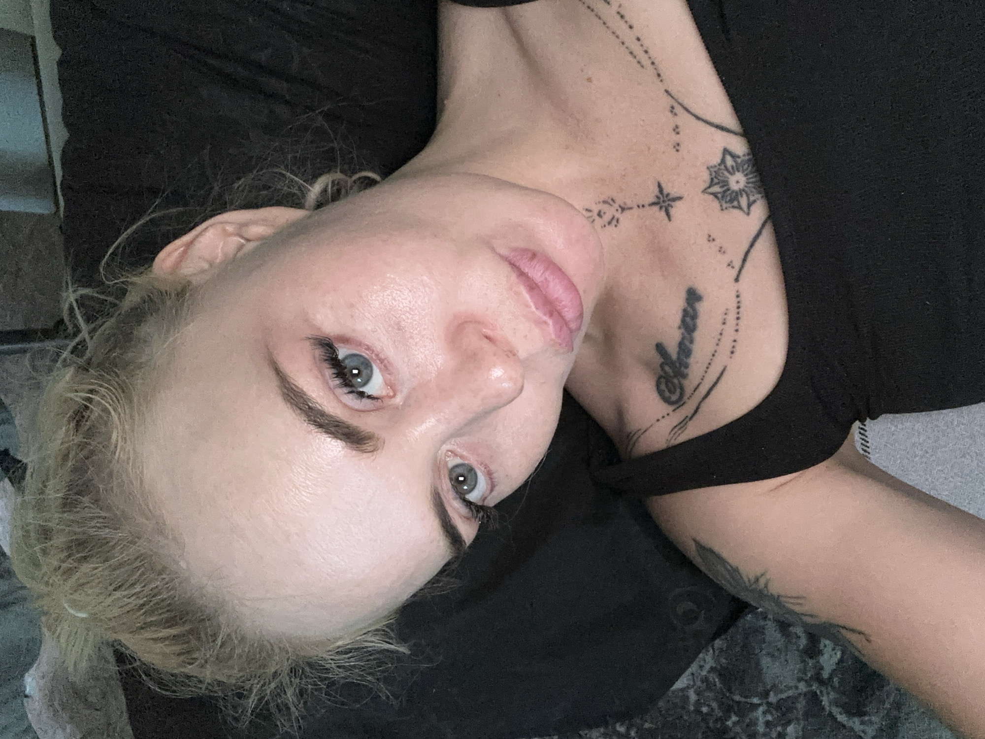 A young woman with blonde hair pulled up in a bun and blue eyes is lying down and looking at the camera. She is wearing a black tank top that shows some of her tattoos on her chest and shoulder. She has makeup on, including mascara and eyeliner. The background is dark and blurry, possibly a bed. The lighting is soft and natural.