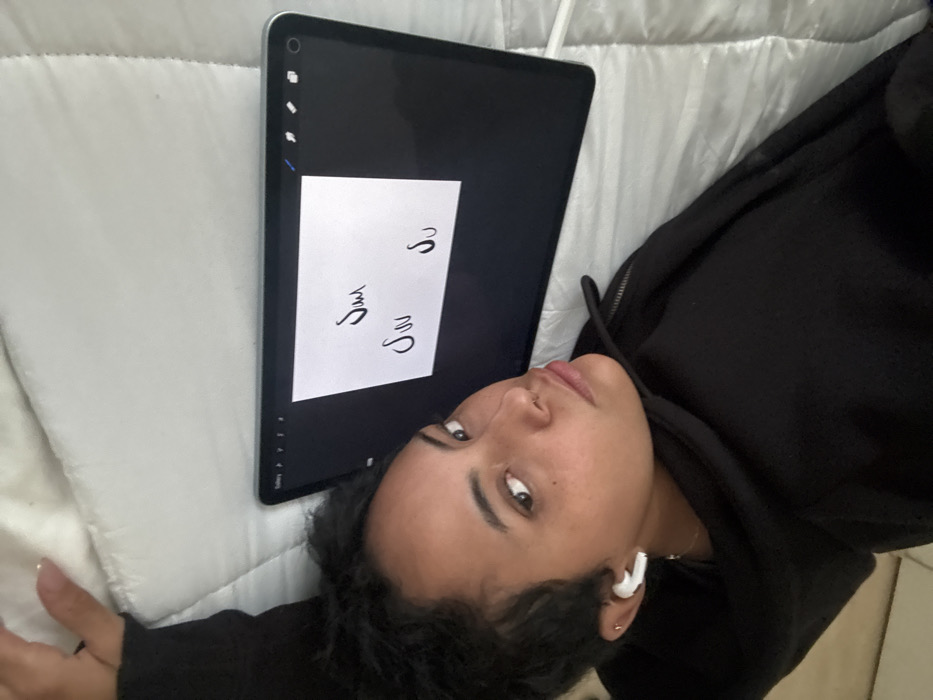 An individual is depicted lying on their back on a light-colored, quilted surface, likely a bed, in an indoor setting. The person, appearing to be a young adult female with dark, curly hair, wears a black hooded sweatshirt and has a white wireless earbud in her right ear, along with a small nose piercing. Her head is turned towards the camera, and she is looking directly into the lens. To her left, an active tablet, displaying a dark interface with a central white digital canvas, is positioned horizontally. Black, stylized script, resembling "ساس" (or three instances of a mirrored 'S' shape), is visible on the white canvas. A white stylus, consistent with an Apple Pencil, rests on the quilted surface near the tablet. The specific location within Mexico City, Mexico, cannot be determined from the image.