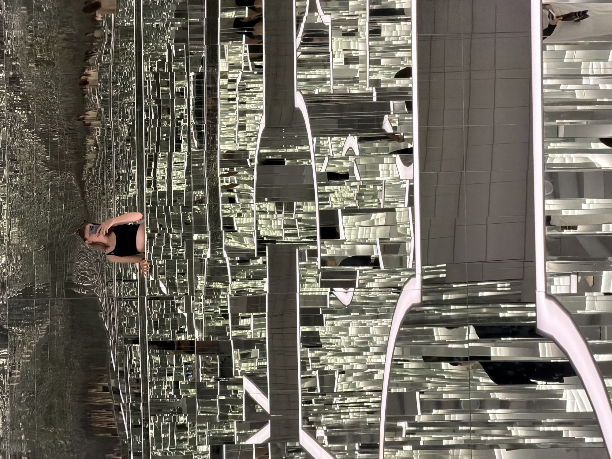 The image depicts a person taking a selfie in a mirrored labyrinth, likely an art installation. The environment is characterized by endless reflections of the individual and the surrounding mirrored structures, creating a disorienting and expansive visual effect. The mirrored walls are arranged in a complex, multi-layered structure with white, illuminated pathways or boundaries. The person, a young woman with auburn hair, is wearing a black crop top and holding a smartphone to take the photo. Several other individuals are visible as reflections in the mirrors, suggesting the presence of other visitors or participants in the installation. The overall impression is one of an immersive, abstract, and visually captivating space. The location is identified as Seoul, South Korea, implying this mirrored labyrinth is a tourist attraction or art exhibit within the city.