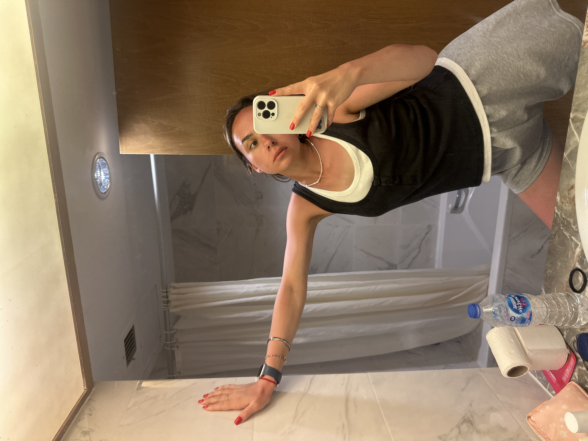 A woman is taking a selfie in a bathroom. She is wearing a black tank top over a white top and grey shorts. Her left hand is resting on a marble countertop and she is holding a white smartphone in her right hand, taking a mirror selfie. Her nails are painted red. There is a bottle of water and a roll of toilet paper on the countertop. A bathtub with a white shower curtain is visible in the mirror. The bathroom has a light fixture in the wall and a wooden-framed mirror. The location is implied to be Pattaya City, Thailand.