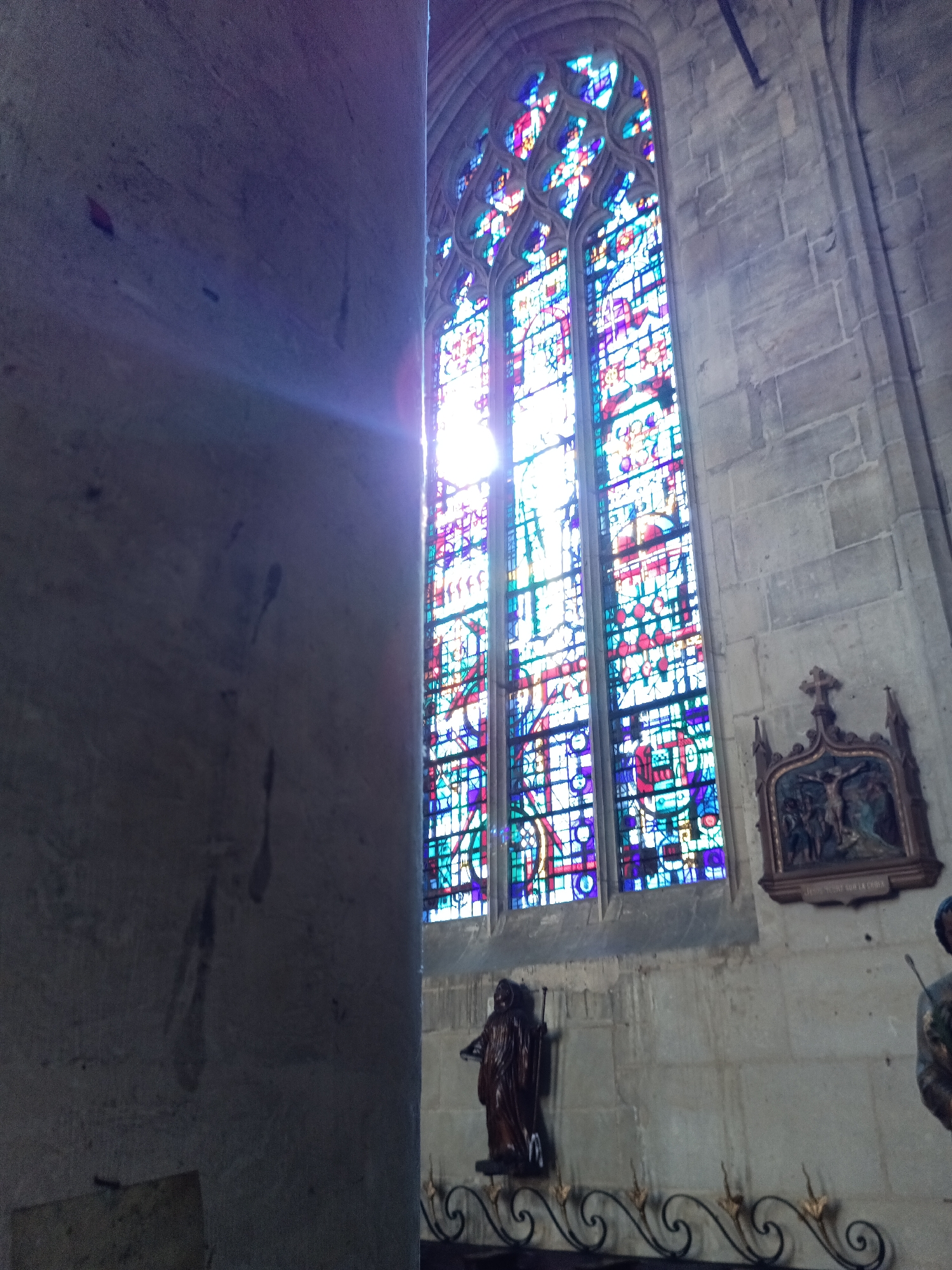 The image shows an interior view of a church in Le Neubourg, France, with a large stained-glass window as the central focus. The window features intricate patterns of colorful glass, primarily blues, reds, and purples, with bright white light shining through it. To the left of the window, a large, rough-textured stone column is prominently visible, with light beams refracting from the stained-glass window onto its surface. The wall behind the window is made of grey stone blocks. Below the window, on the right side, there is a wooden decorative panel with a crucifixion scene depicting Jesus on the cross, with figures of Mary and John beside him. Further to the right, a dark, silhouetted statue of a figure in robes stands on a pedestal, and another statue, partially visible on the far right, also appears to be a religious figure. A decorative metal railing with swirling patterns runs along the bottom of the scene, in front of the statues. The overall impression is one of a dimly lit, historic church interior, with the stained-glass window and the religious artifacts being the primary subjects.