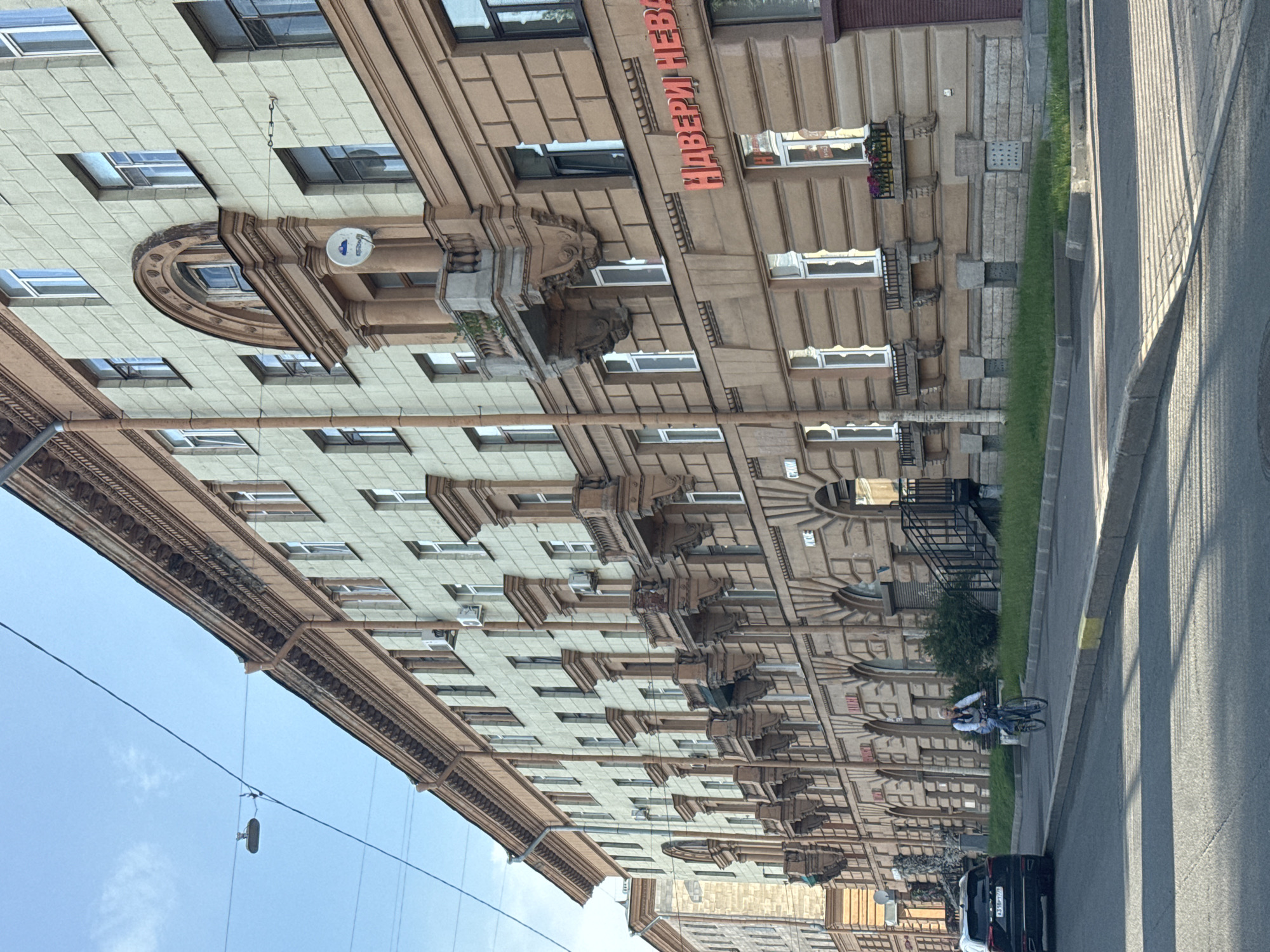 The image depicts a wide, sunlit street in Saint Petersburg, Russia, with a long, light-green and brown building lining the right side. The building has a classical architectural style with repeating windows, balconies with ornate supports, and decorative cornices. A red neon sign with Russian text "НАВЕРИ НЕВА" is visible on the building's facade.
In the foreground, the street surface is asphalt, with distinct shadows cast by an unseen object to the left. A bicycle with a person riding it is positioned on the sidewalk to the right of the street, moving away from the viewer. The person is wearing a light-colored shirt and dark pants. To the far left of the image, the rear of a dark SUV is visible, parked along the side of the road.
The sky above is a clear blue with a few wisps of white clouds. Electrical wires are strung across the upper portion of the frame. The overall scene suggests a typical urban environment in Saint Petersburg on a bright, clear day.