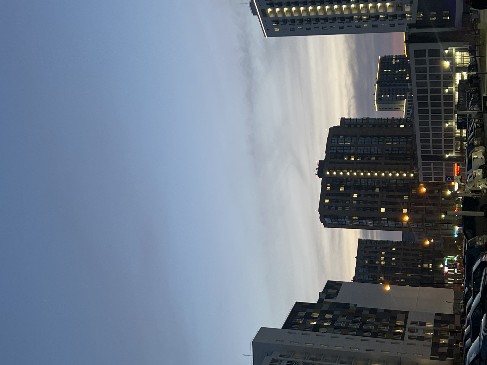 The image captures an urban scene in Tyumen, Russia, at dusk or dawn. The sky transitions from a deep blue on the left to lighter shades with hints of pink and orange near the horizon, where the cityscape meets the sky. Several high-rise buildings dominate the view, their windows illuminated. The buildings vary in height and architectural style. A parking lot is situated on the right. Cars of various makes and models are densely packed together, suggesting a busy urban environment. Streetlights and other illuminated signs add to the light. The overall impression is one of a modern urban setting, likely a residential or commercial district.