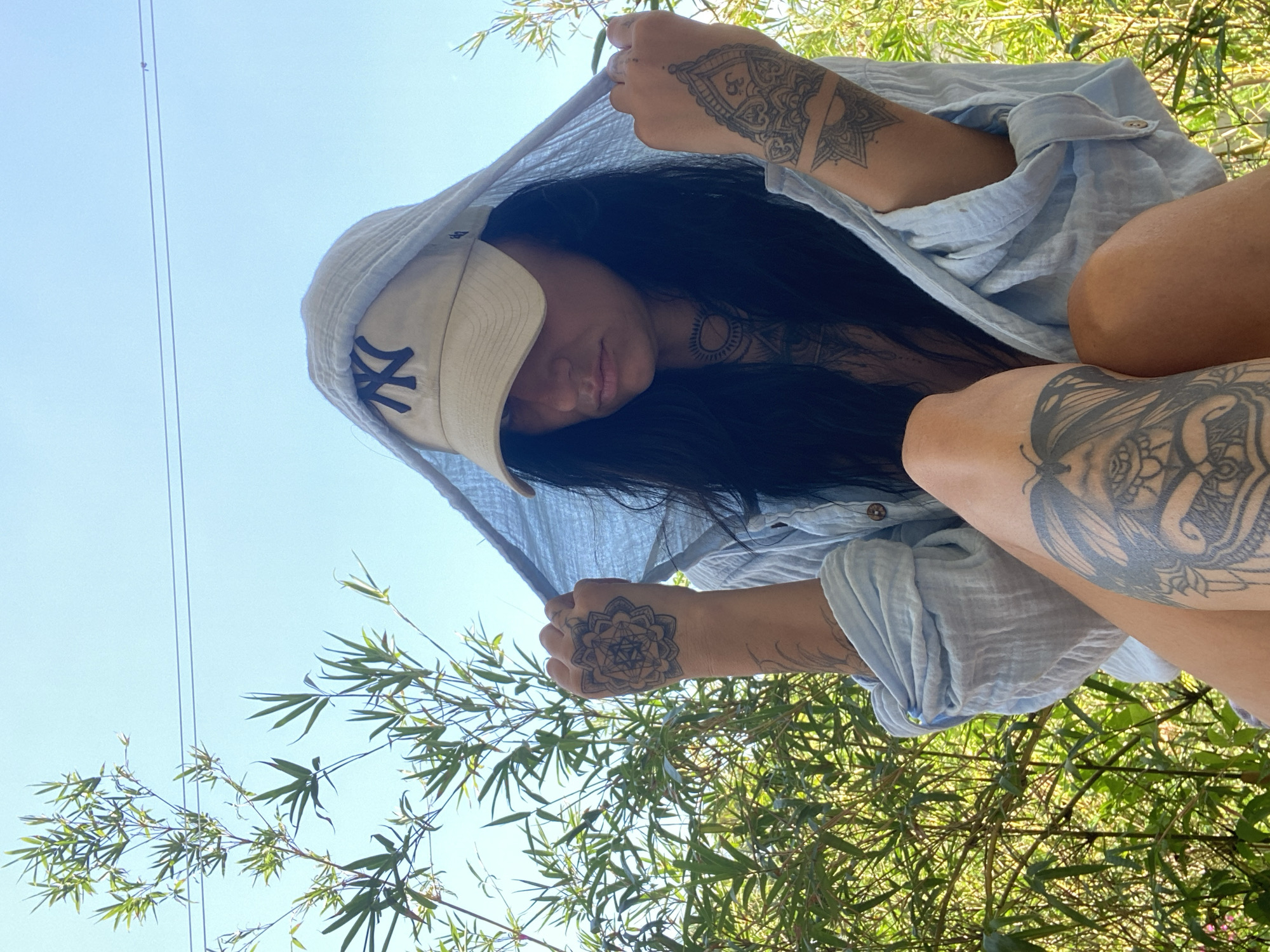 A young woman is photographed from a low angle. She wears a baseball cap with a "NY" logo and a light blue button-up shirt. The shirt is draped over her head as a makeshift hood. Both arms are visible, and each has elaborate tattoo designs. The woman appears to be outdoors, surrounded by green foliage, possibly bamboo. Power lines are visible against a bright blue sky, suggesting an urban or semi-urban location. Based on the request, this likely took place in Mũi Né, Vietnam.