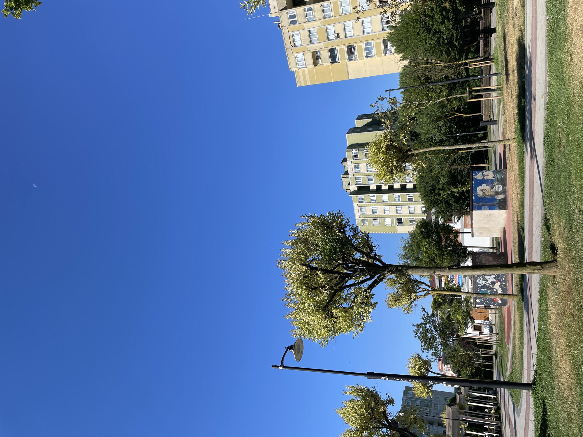This is a photograph of a public park in Başakşehir, Turkey, under a clear blue sky with a faint crescent moon visible. The park features paved walkways with red and grey borders, green grass, and several trees, some of which have light-colored foliage. A black lamppost stands on the left side of the image. In the background, there are several multi-story apartment buildings, one in a light yellow color and another in a greenish hue. There are also some graffiti-covered structures and park benches visible within the park. The overall scene depicts a quiet, daytime urban environment.